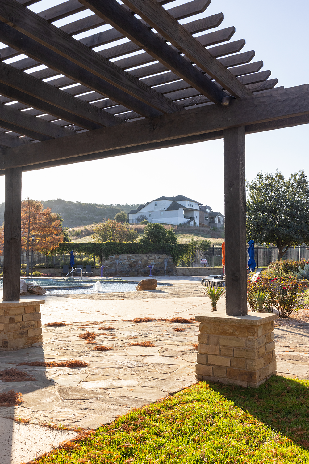 View of a swimming pool and patio seating under a pergola, with a house on a hillside in the background.