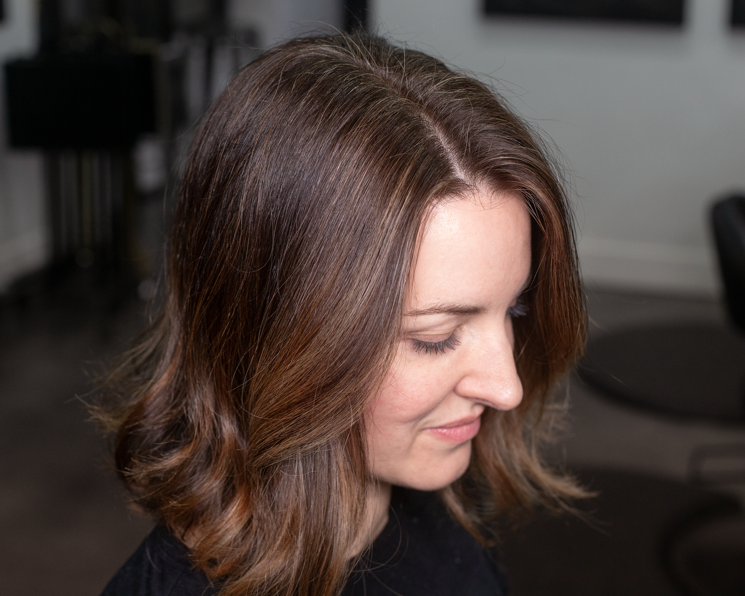 Close-up of a woman's head with shoulder-length brown hair styled in loose waves, looking downward in an indoor setting.