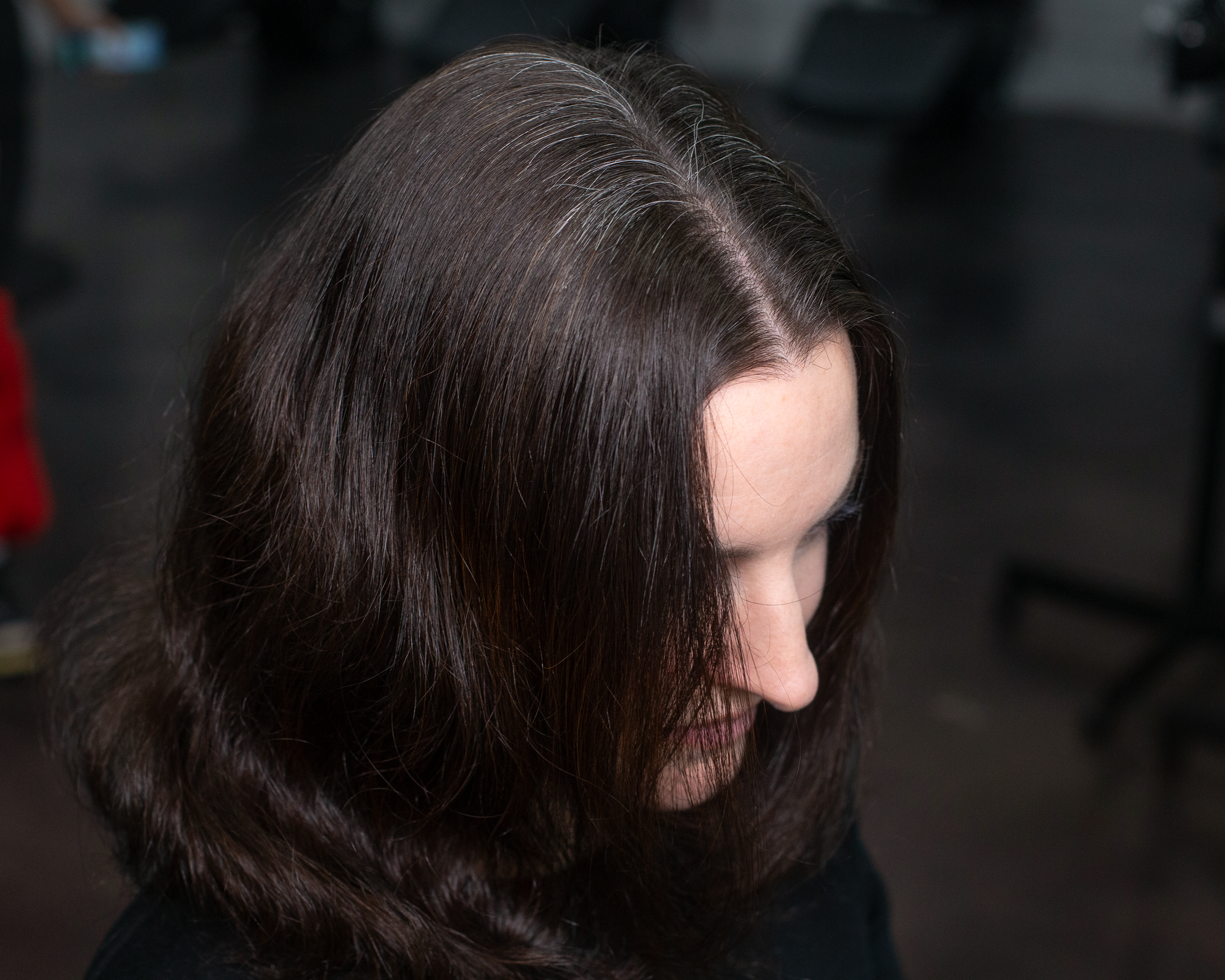 A woman with shoulder-length dark brown hair with gray roots, looking downward with her face slightly turned to the side.