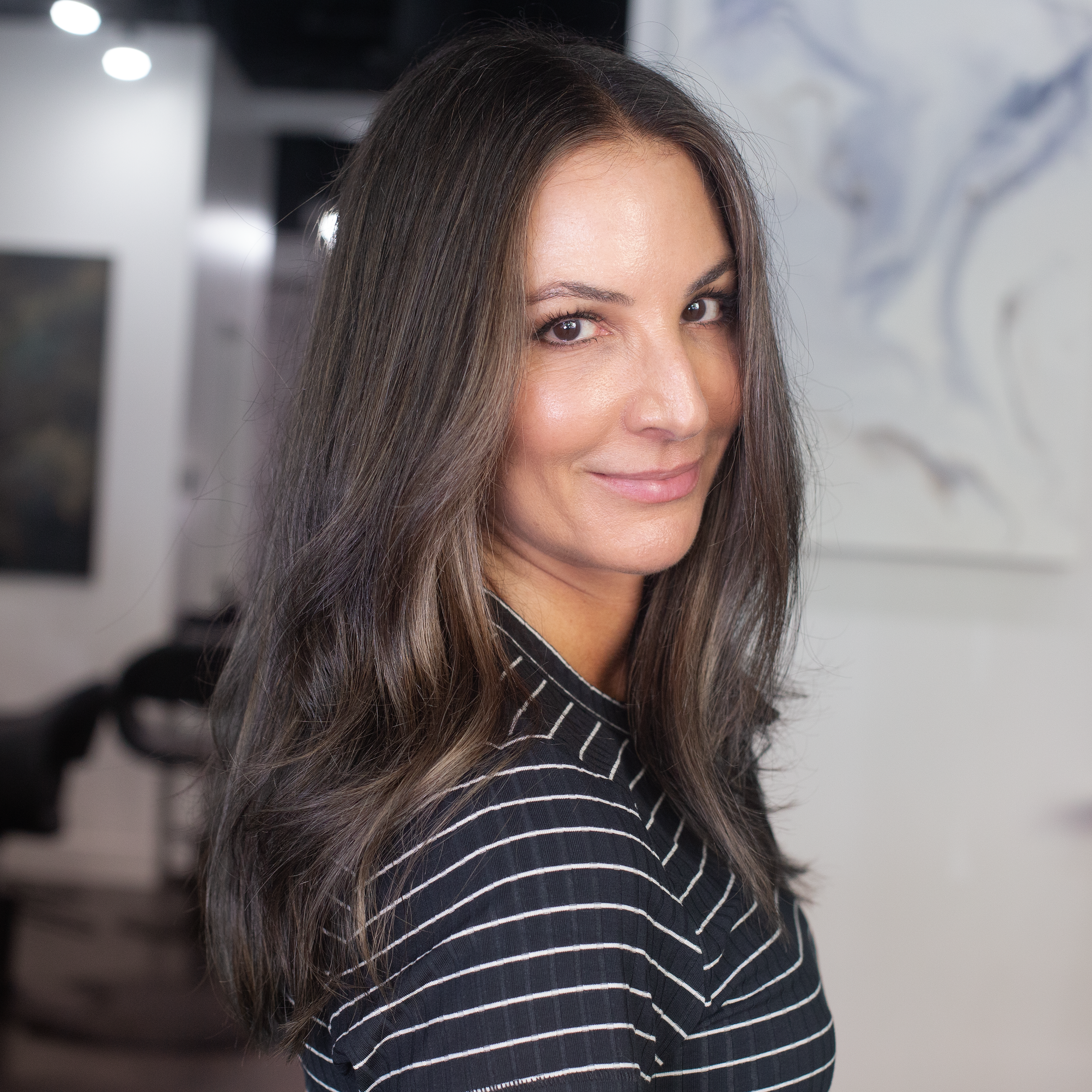 A woman with long, layered grayish brown hair and subtle makeup, smiling softly, wearing a black and white striped shirt, standing in an art gallery with artworks on the wall behind her.