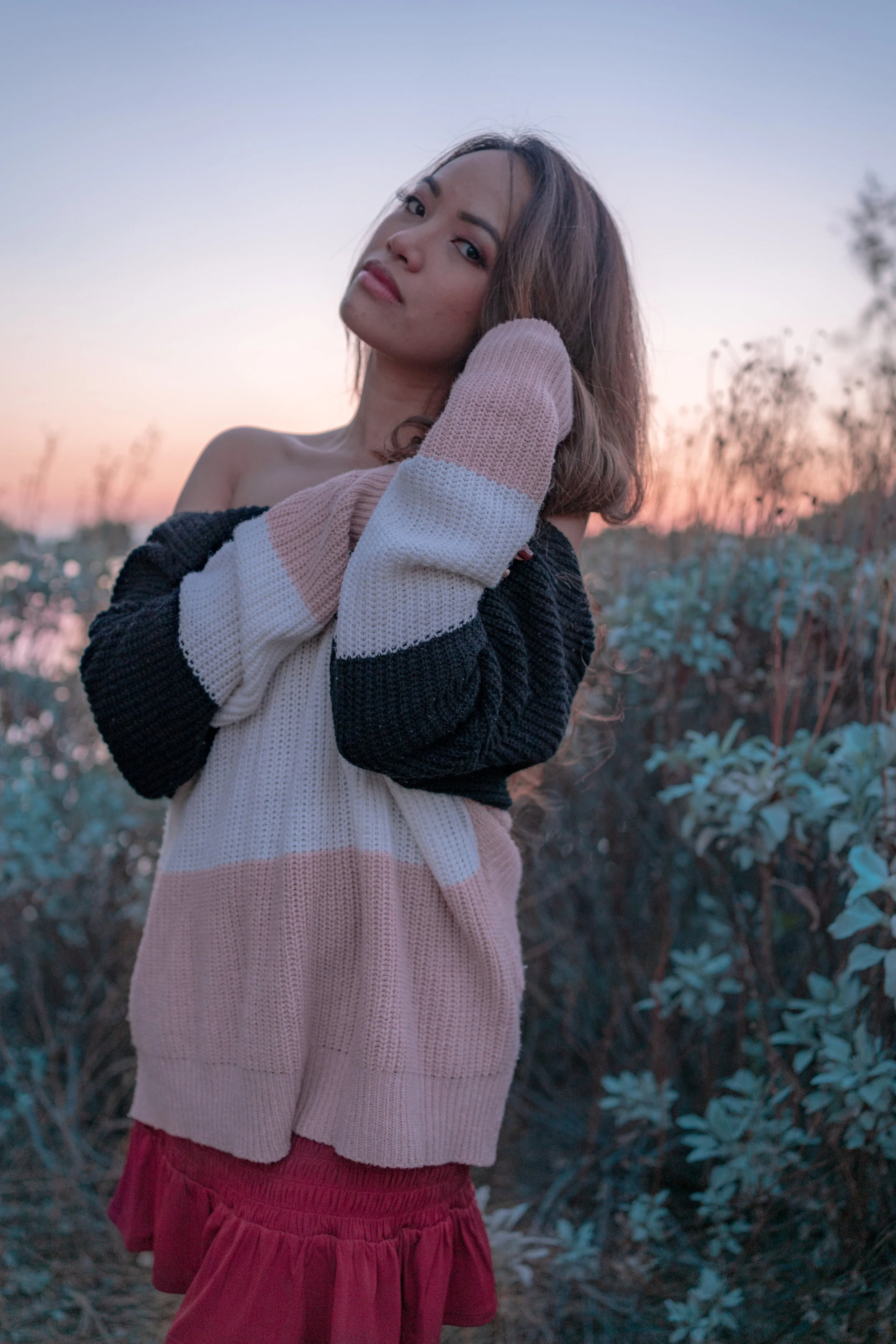 A woman with shoulder-length brown hair, wearing a striped knit sweater and a red skirt, standing outdoors at sunset, with a calm expression and her hand resting behind her neck.