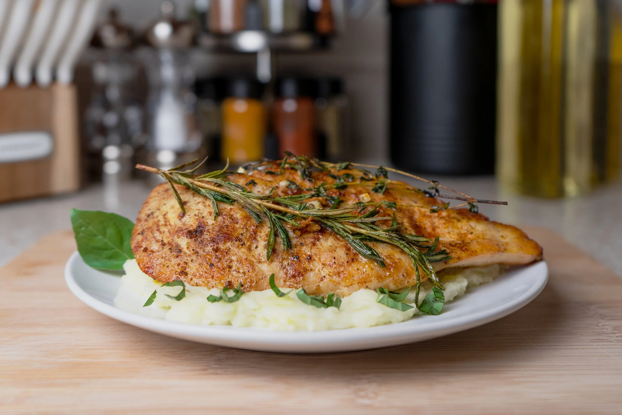 Cooked chicken breast topped with rosemary and herbs, served on mashed potatoes with green herbs on the side in a white dish on a wooden surface.