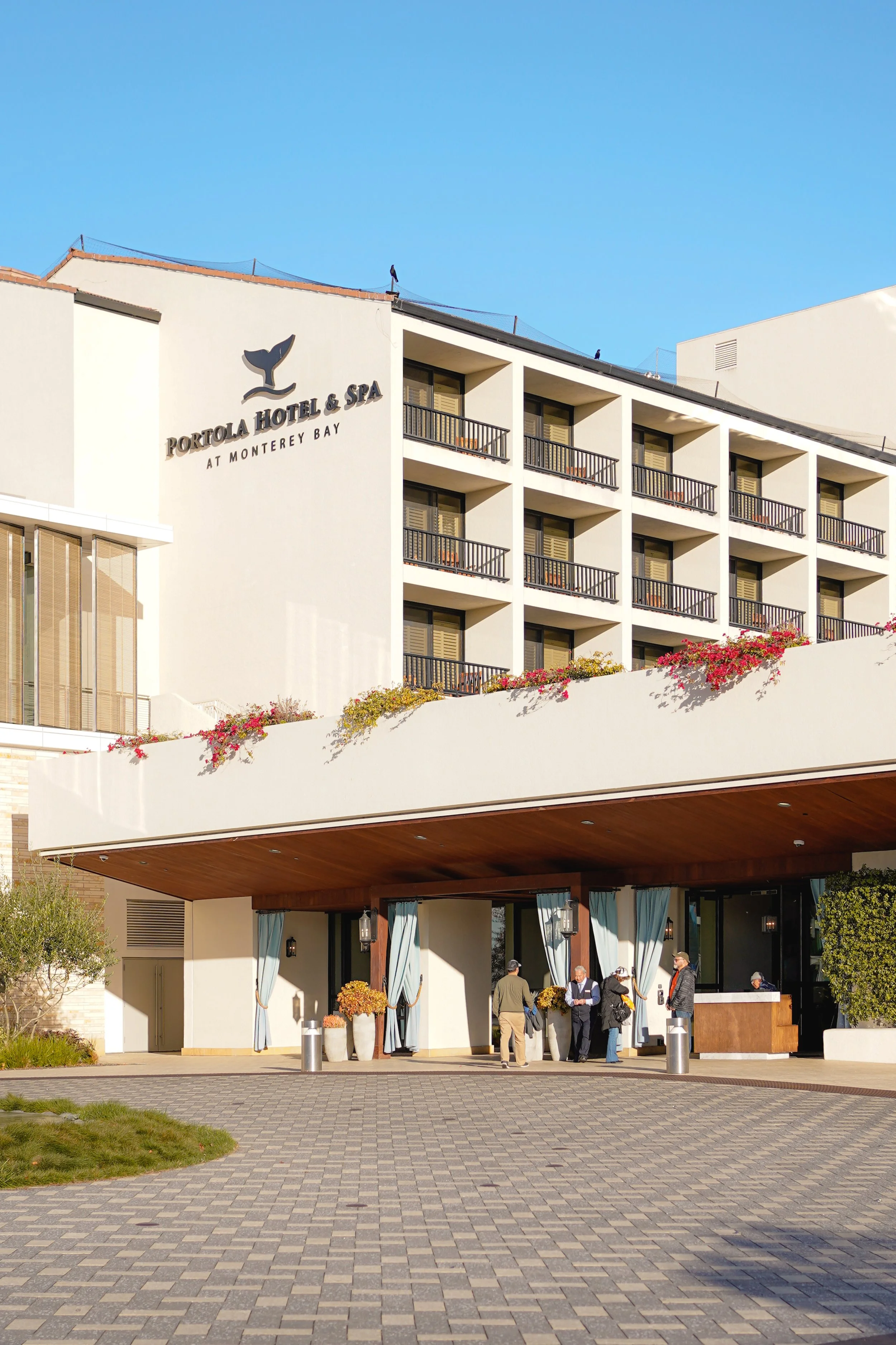The exterior of Portola Hotel & Spa at Monterey Bay with guests checking in at the entrance, under a covered area, with multiple balcony rooms above and the hotel logo on the wall.