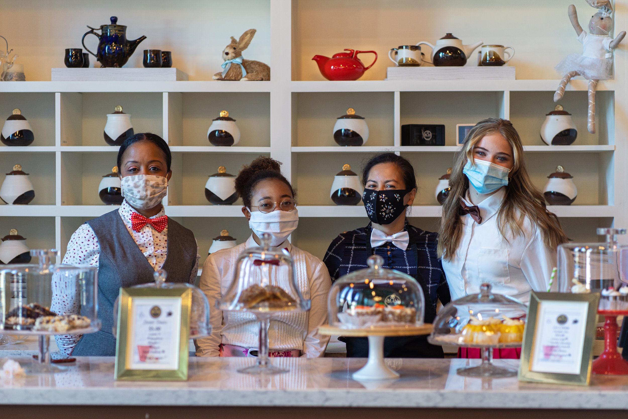 Four women wearing face masks behind a table full of desserts at a bakery or cafe. Shelves with teapots and decorative items in the background.