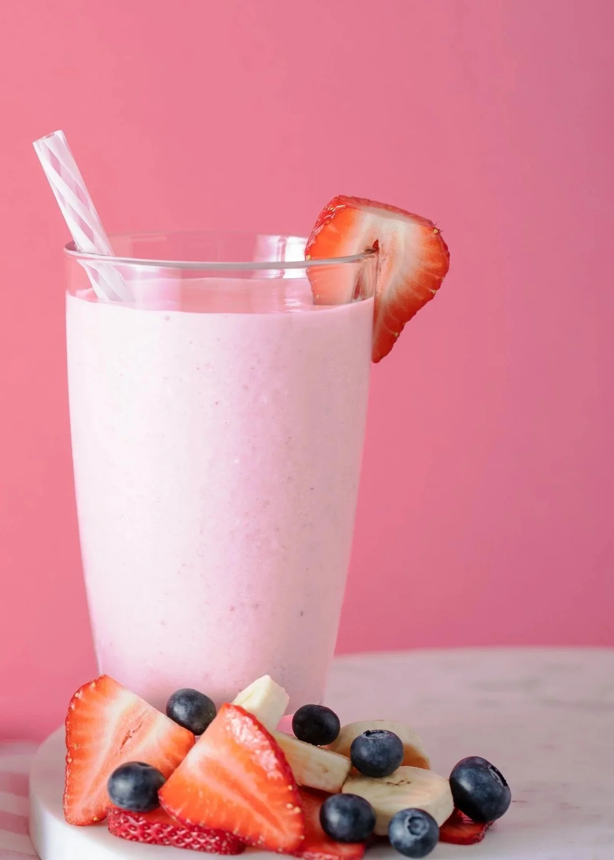 A pink strawberry and blueberry smoothie in a tall glass with a striped straw, garnished with a strawberry slice on the rim, with sliced strawberries, blueberries, and banana on a white plate in front, against a pink background.