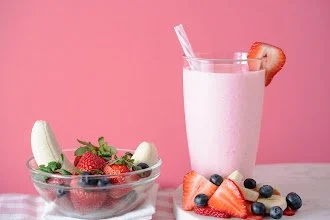 A glass of pink strawberry milkshake with a straw and a strawberry garnish, a bowl of mixed berries including strawberries, blueberries, and cream, and some sliced strawberries and blueberries on a pink background.