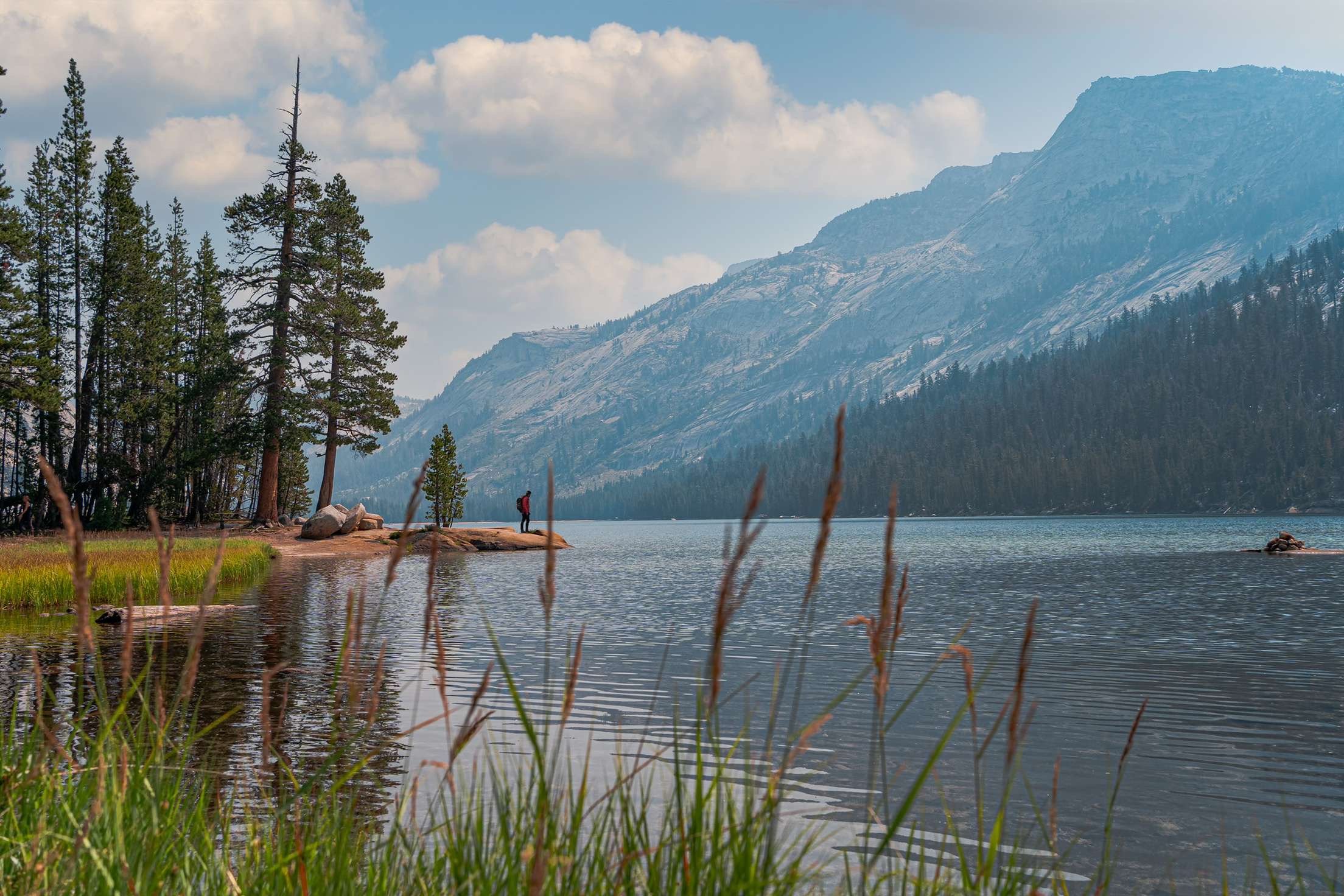 A person standing on a rock by a lake, surrounded by tall pine trees and mountains under a partly cloudy sky.
