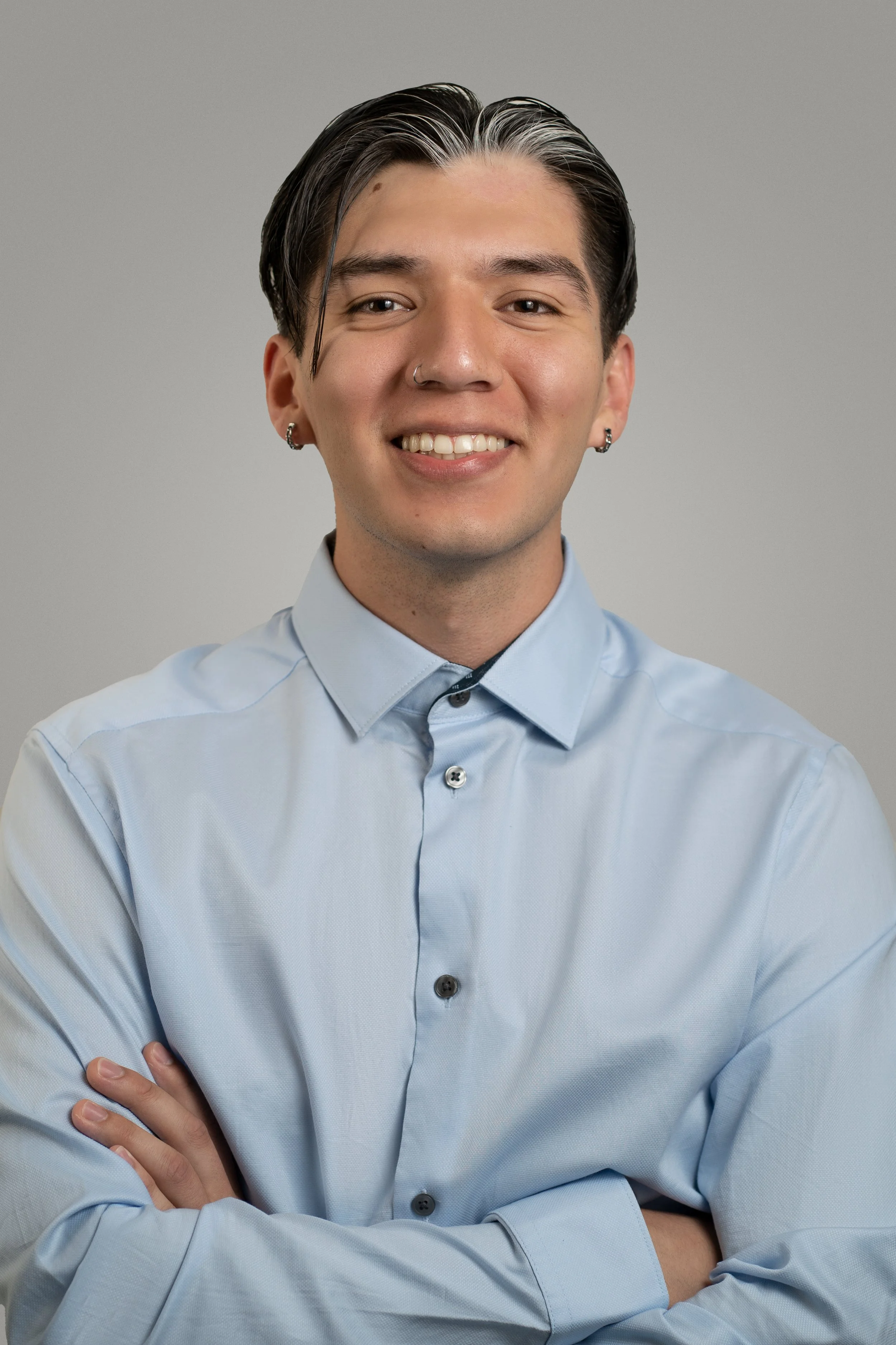 A person with short dark hair, wearing a light blue button-up shirt, smiling, with a gray background.