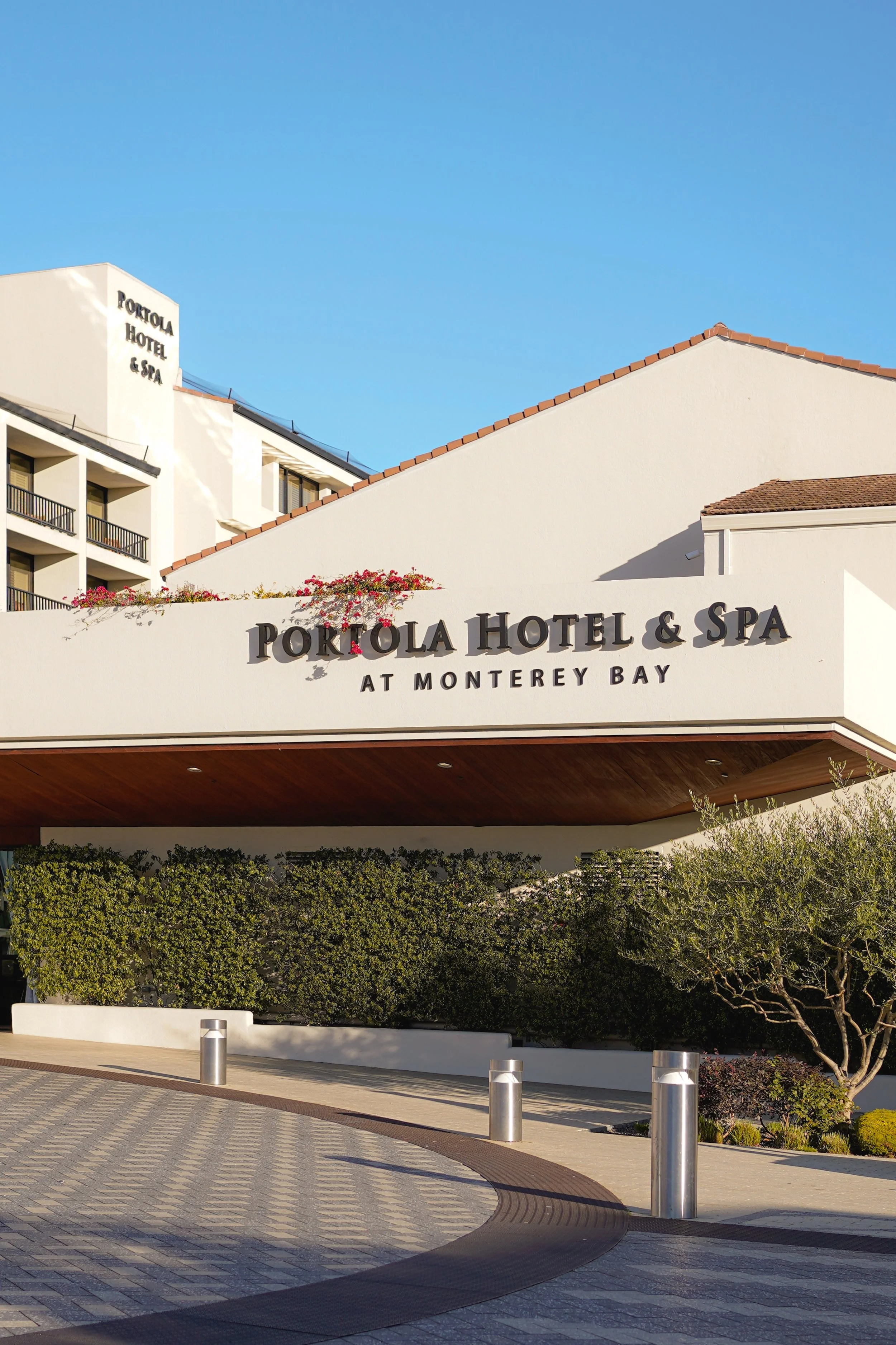 The entrance of the Portola Hotel & Spa at Monterey Bay with white building walls, red-tiled roof, greenery, and clear blue sky.