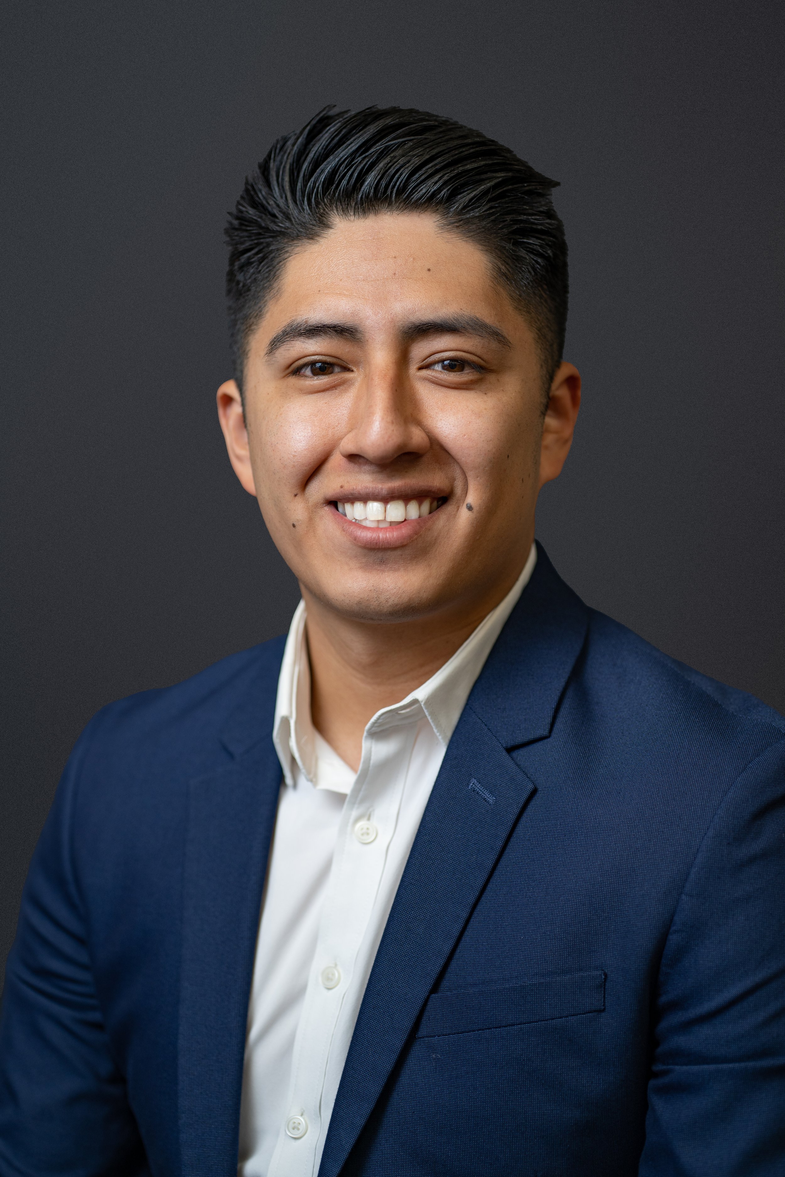 Portrait of a young man with black hair styled neatly, wearing a white shirt and a navy blue blazer, smiling against a dark gray background.