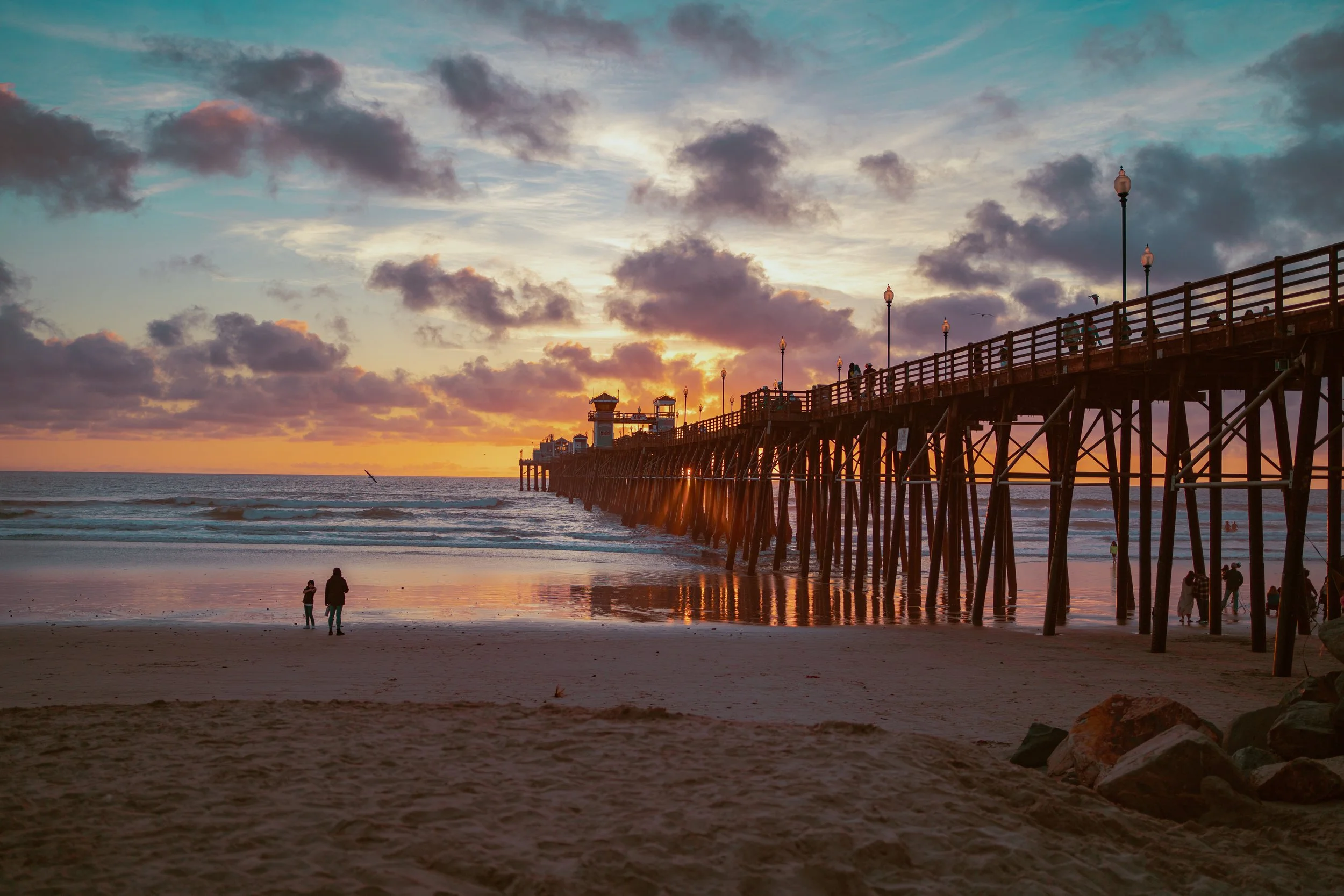 Sunset at the beach with a pier extending into the water, people walking along the shore, and a sky filled with clouds.