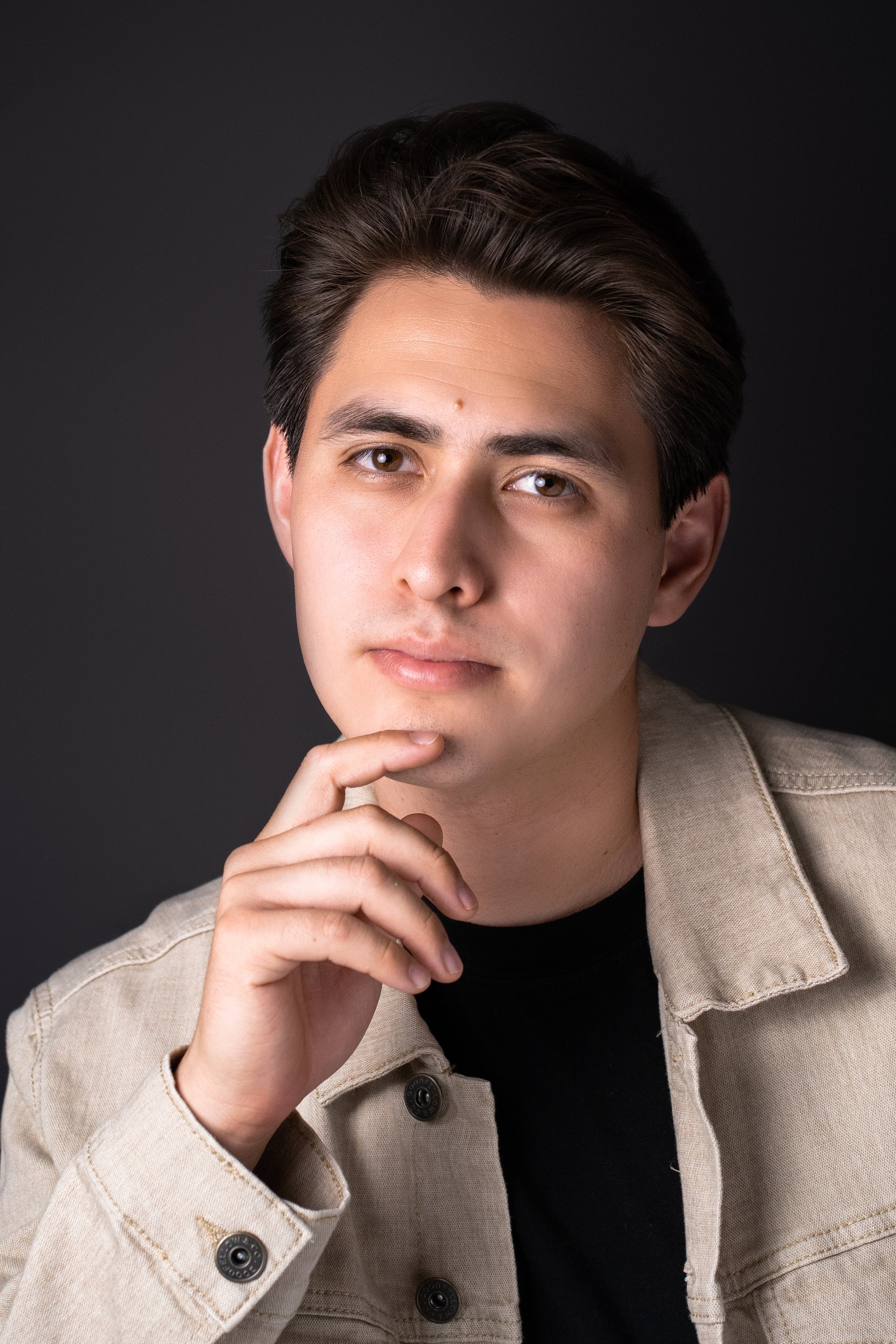 Portrait of a young man with neatly styled dark hair, wearing a beige jacket over a black t-shirt, touching his chin with his right hand, against a dark gray background.