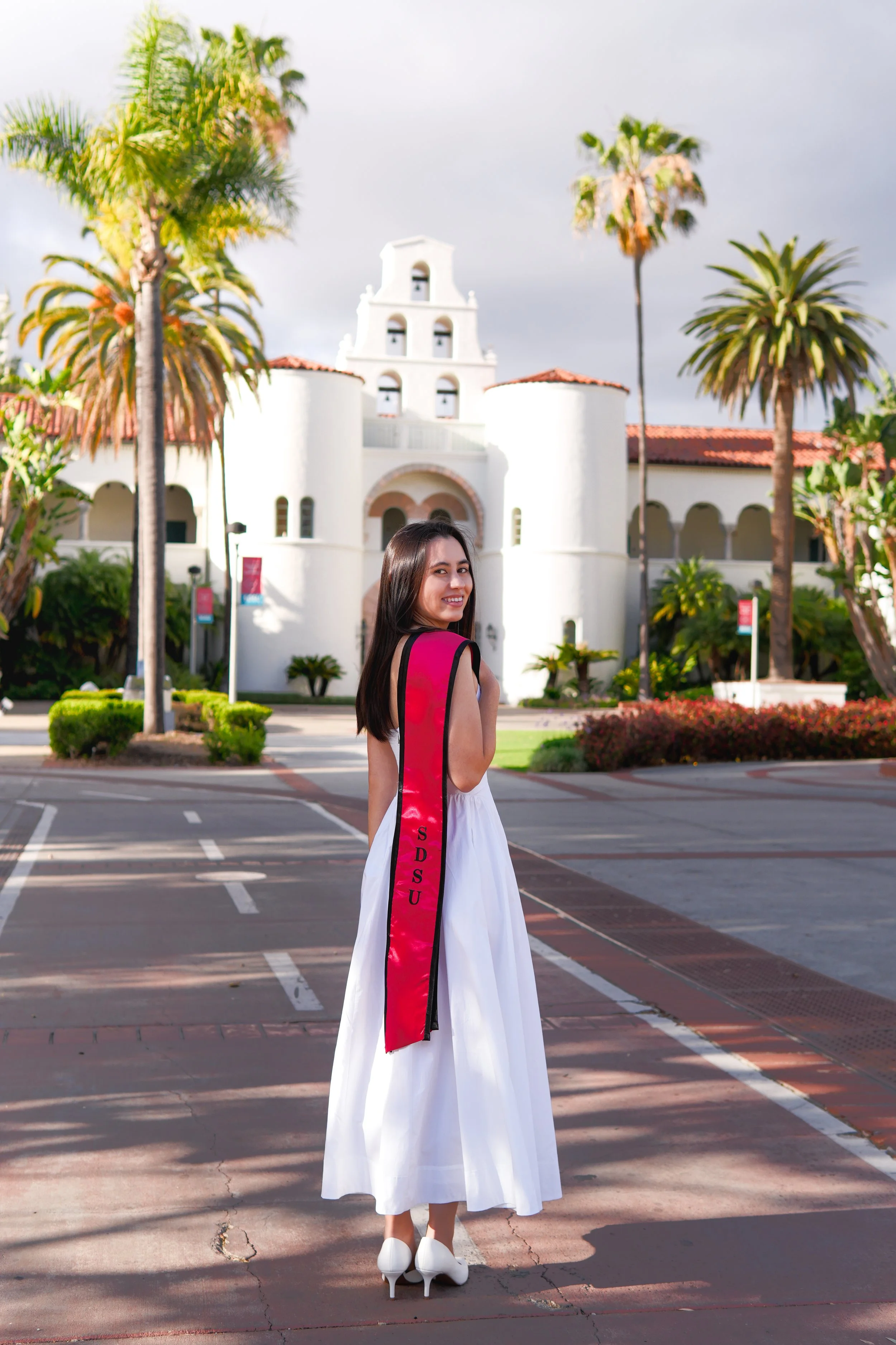 A young woman in a white dress and high heels standing outdoors in front of a historic white building with red-tiled roofs and palm trees, smiling at the camera.