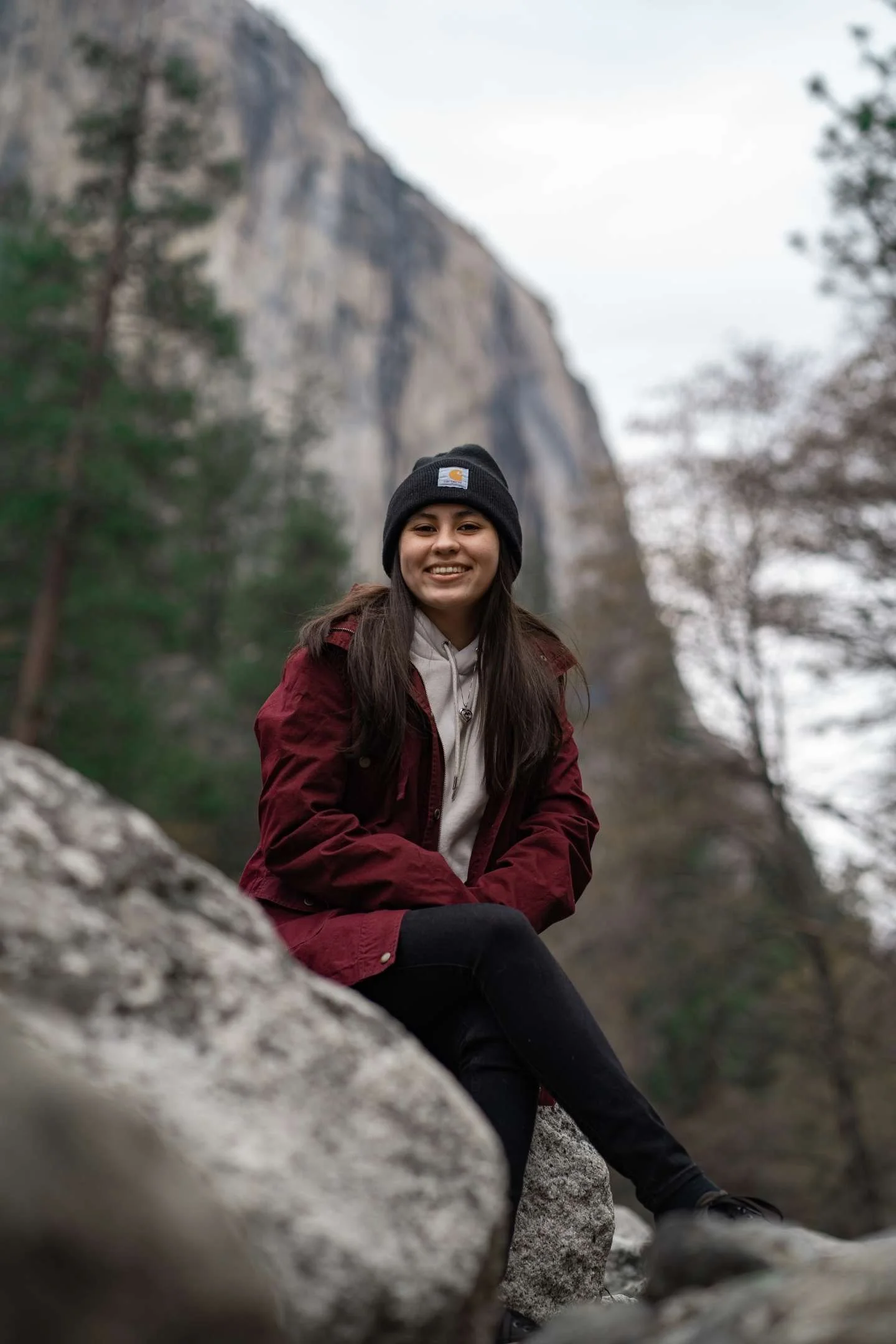 Young woman sitting on rocks outdoors with a mountain and trees in the background, smiling at the camera.