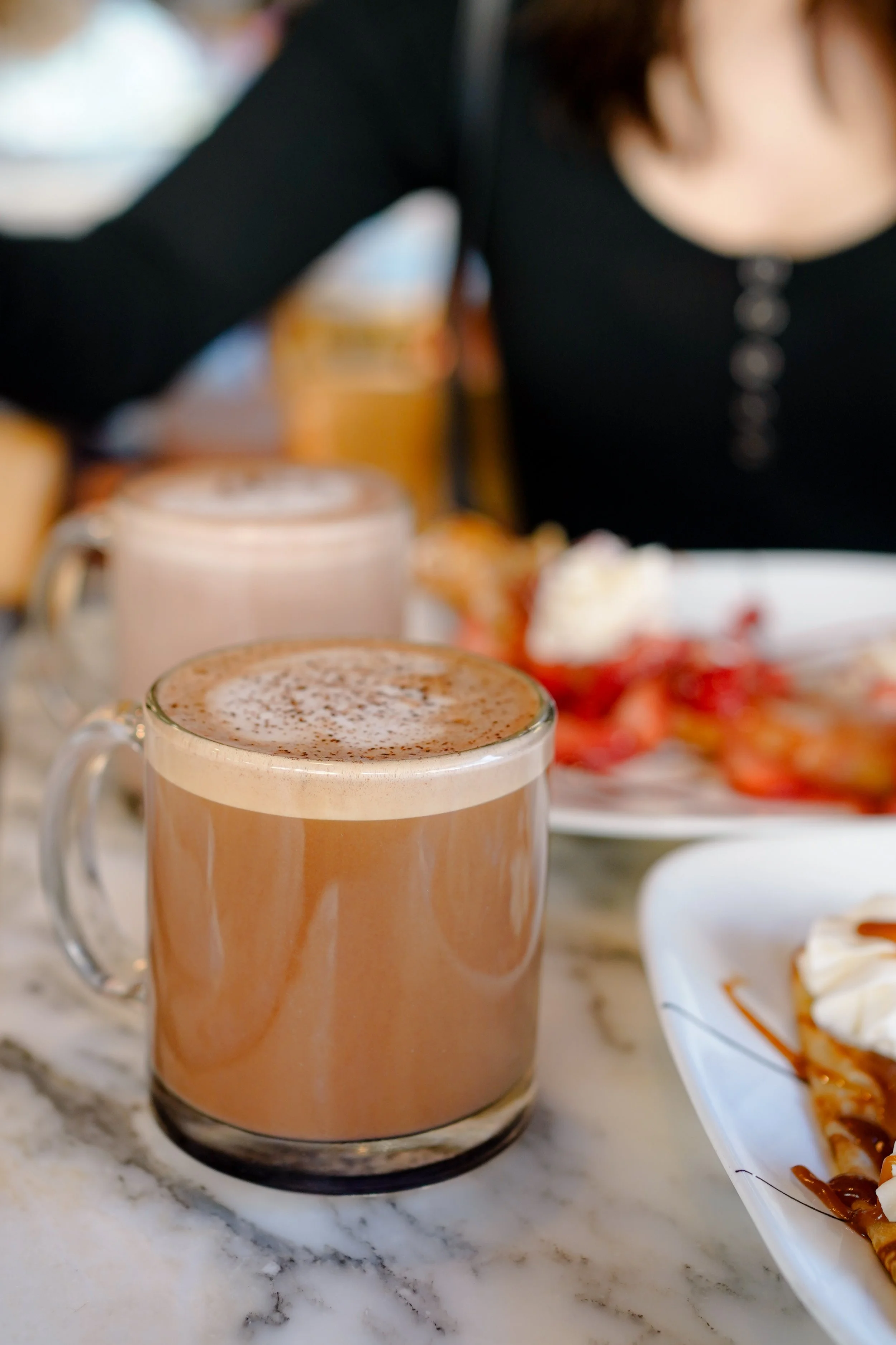 A clear glass mug filled with a hot chocolate beverage topped with whipped cream and cocoa powder, placed on a white marble surface with a plate of strawberries and a dessert in the background.