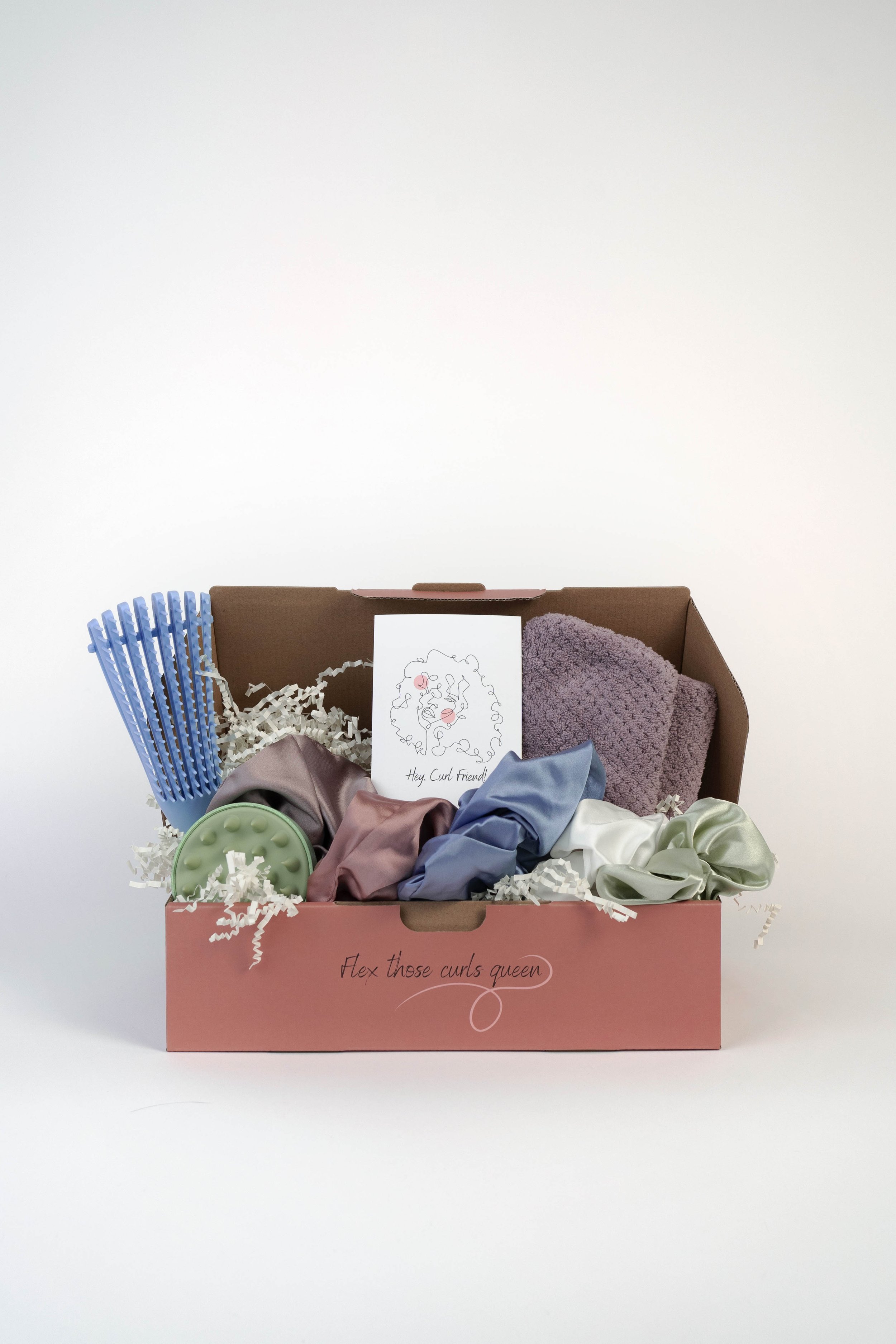 Pink gift box labeled "Flex those curls queen" filled with hair accessories including scrunchies, headbands, and a ring light, with white shredded paper and a greeting card inside.