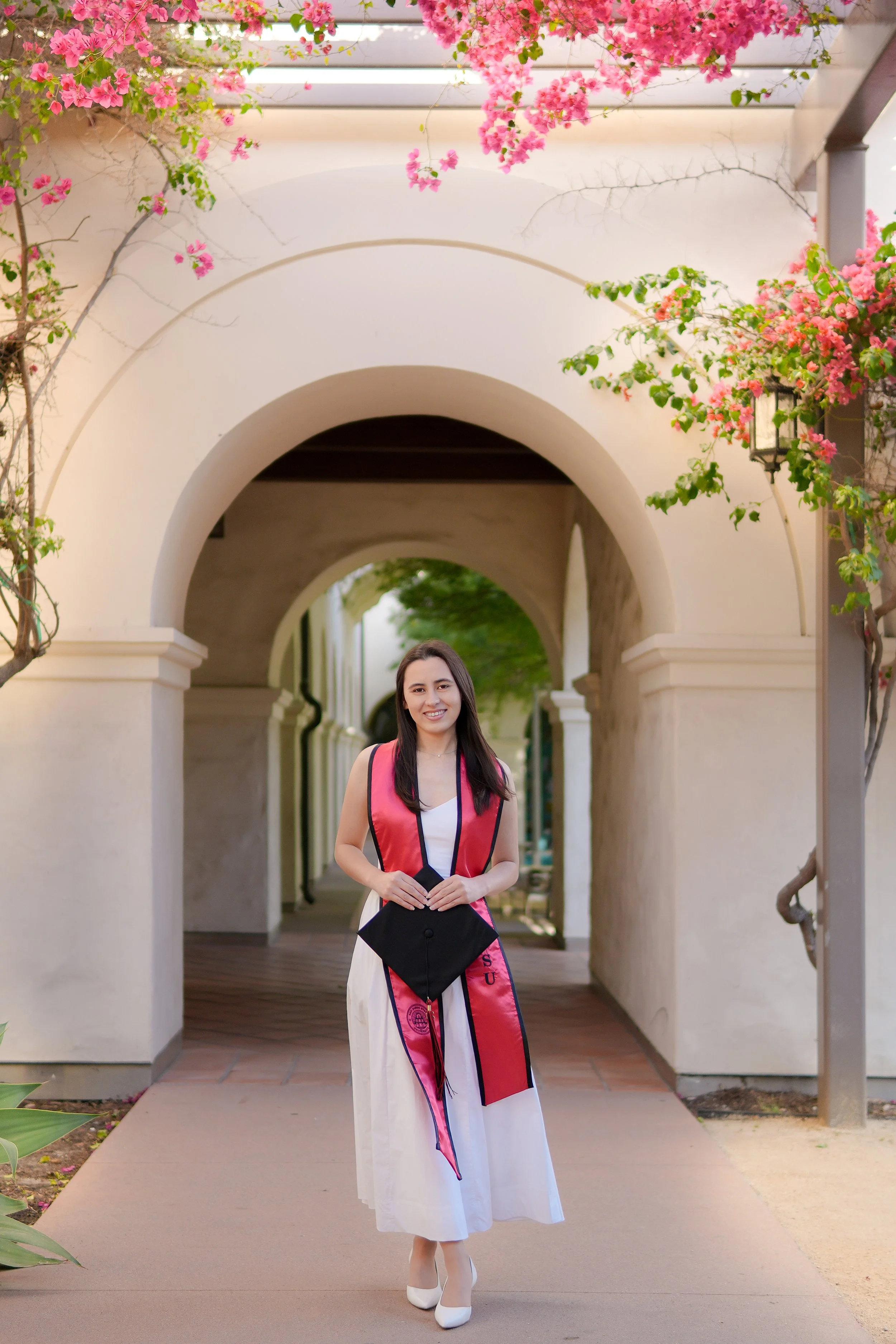 A young woman in a white dress and pink graduation stole stands outside under a white archway with pink flowers.
