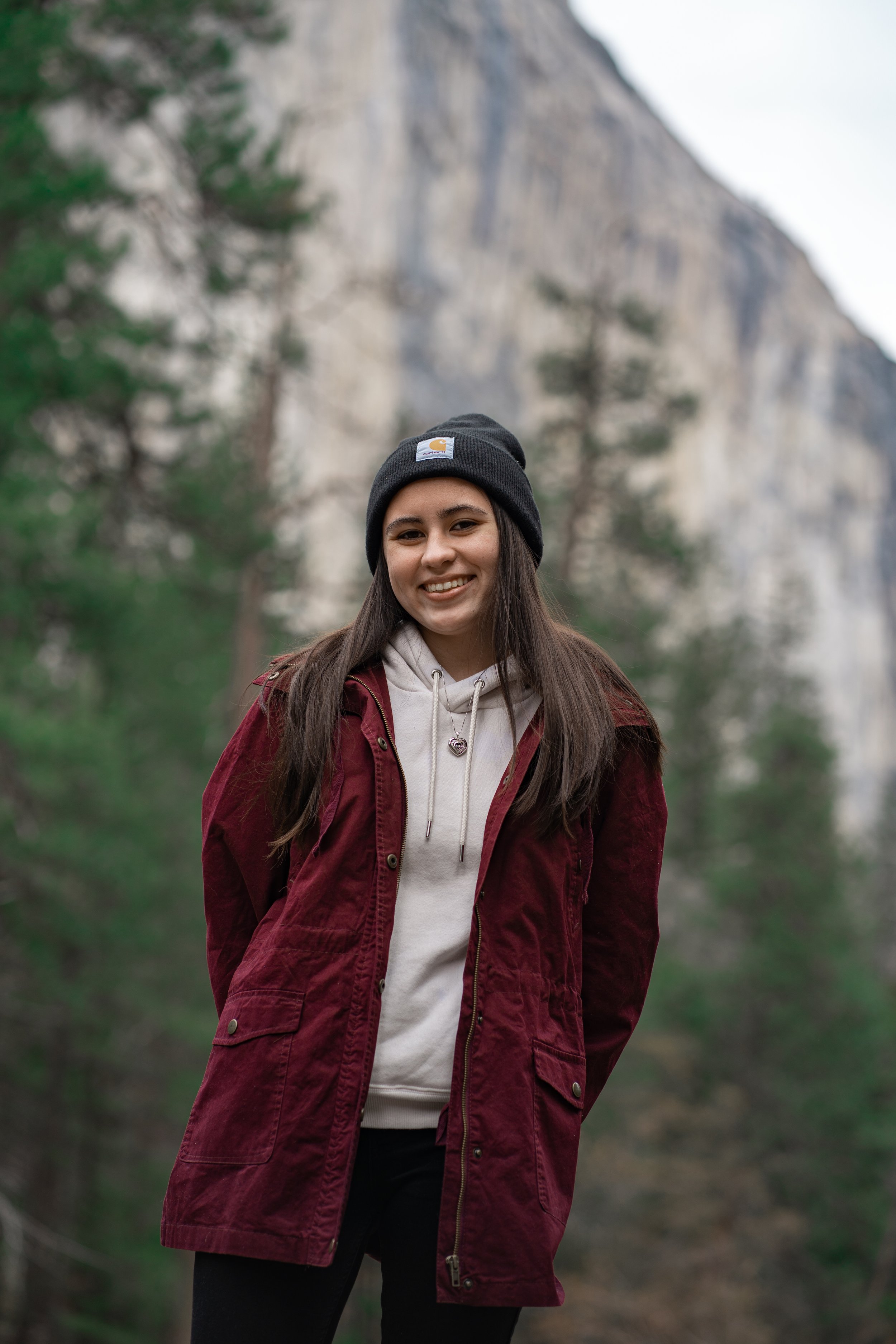 Young woman outdoors in natural setting with trees and cliffs, wearing a black beanie, beige hoodie, and red jacket.