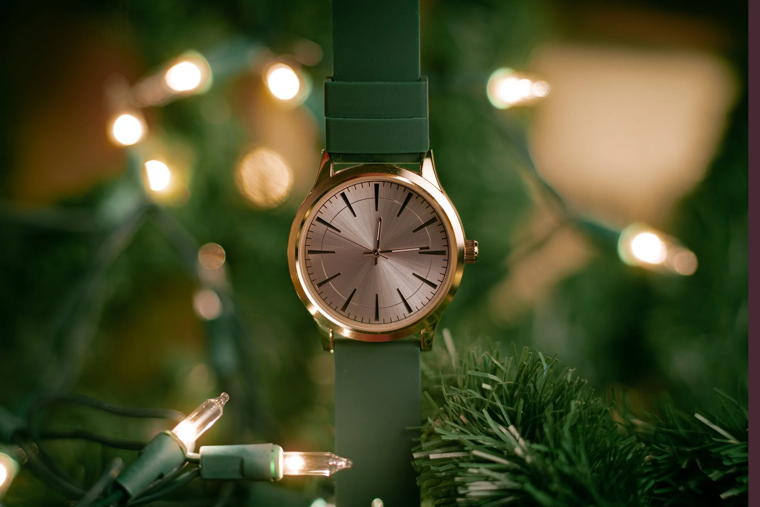 A wristwatch with a gold case and neutral face hanging from Christmas tree lights, surrounded by green branches and out-of-focus lights.
