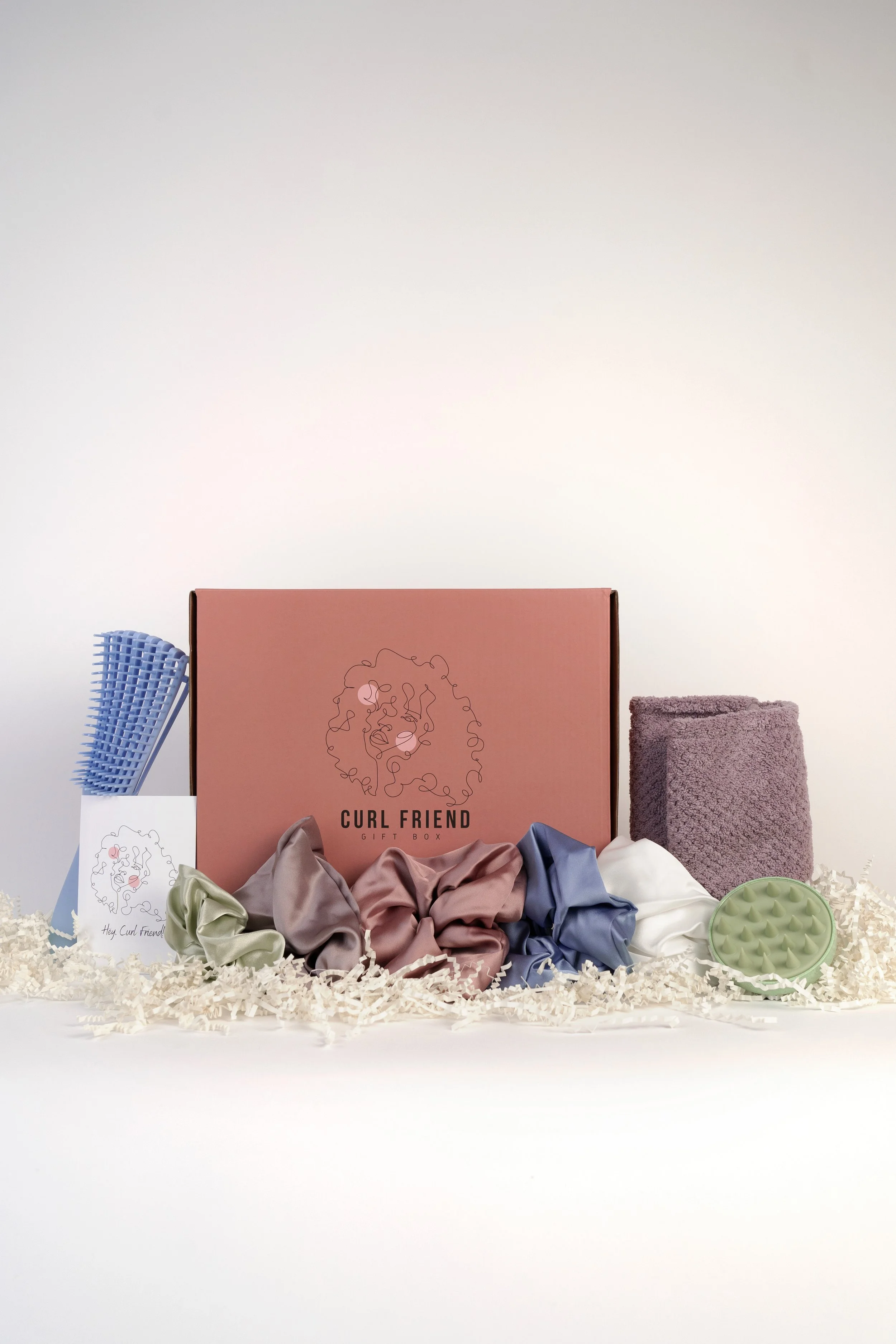 A pink box labeled 'Curl Friend Gift Box' surrounded by scrunchies, paper, a brush, a towel, a silicone scalp massager, and shredded paper on a white background.