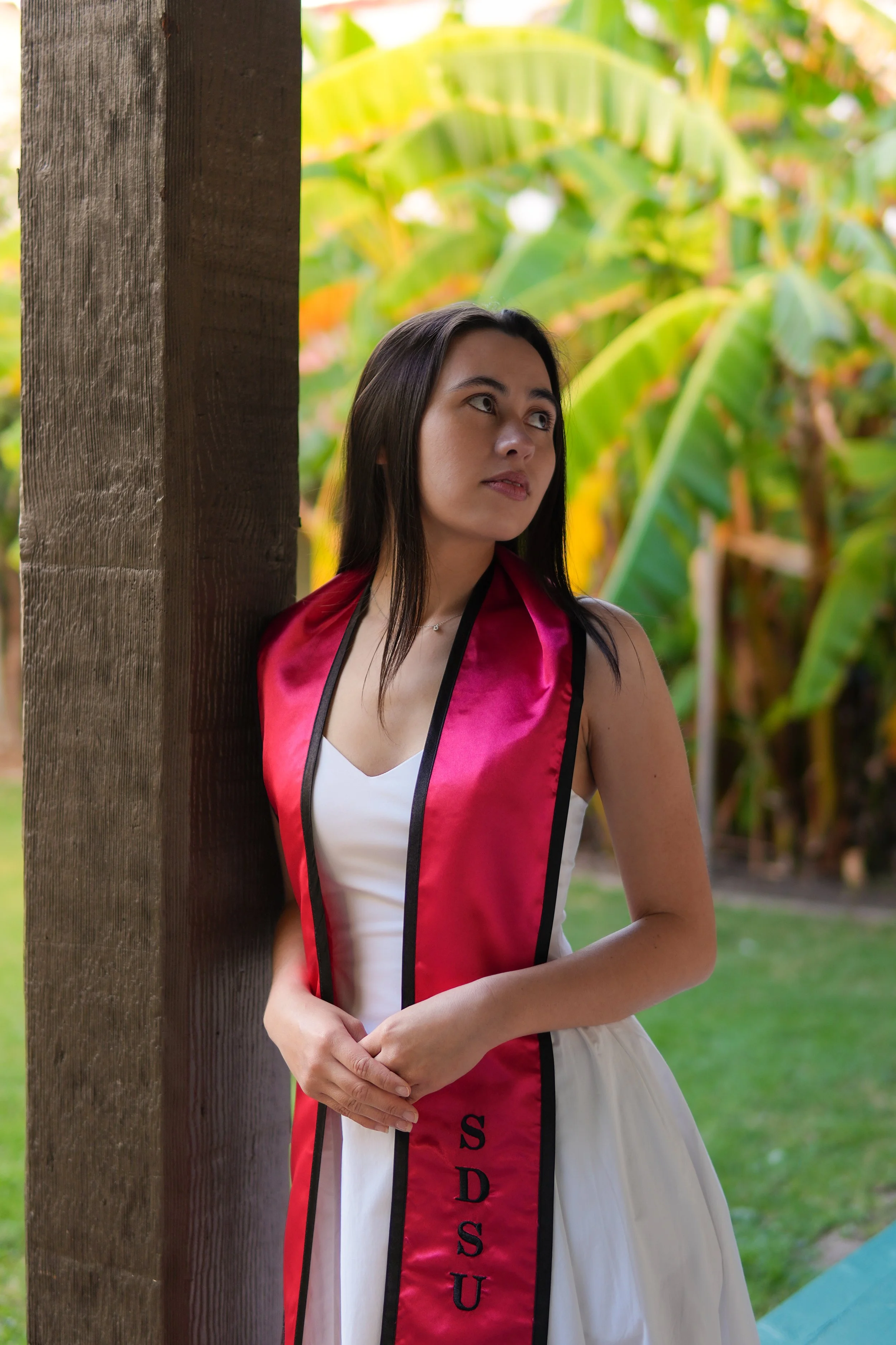 A woman wearing a white dress and a red sash with black trim and the word 'SDSU' printed vertically on it, standing outdoors beside a wooden post with a background of green banana leaves.