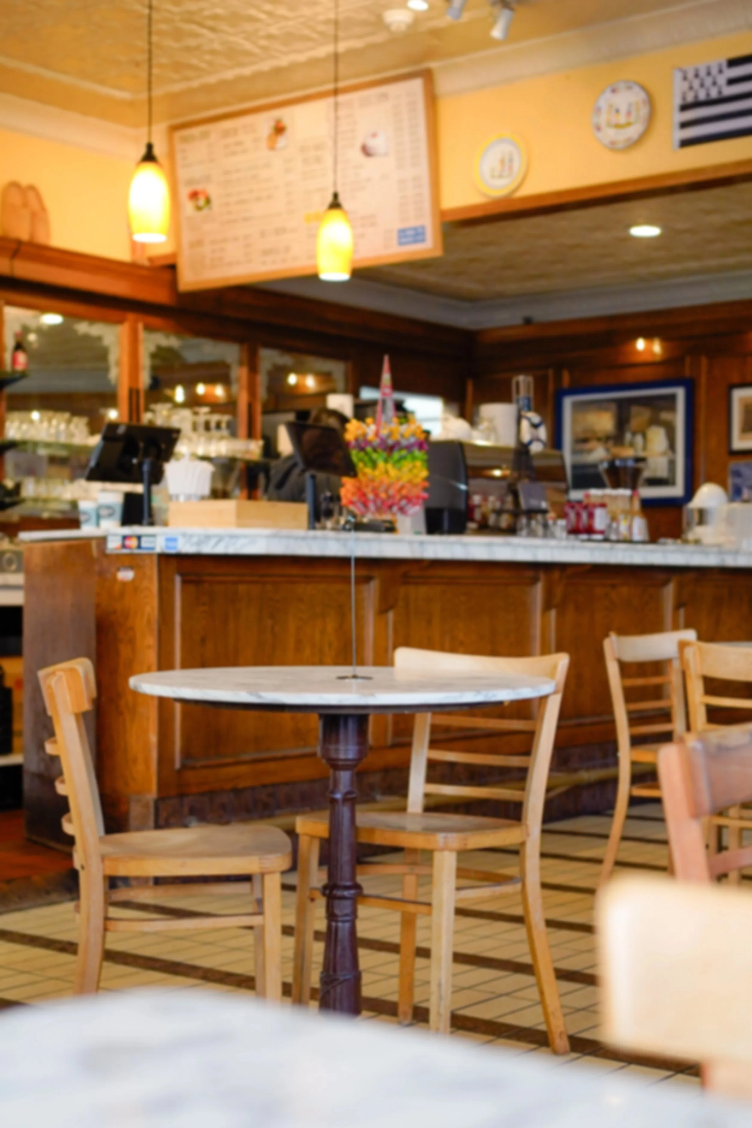 Interior of a restaurant or cafe with wooden furniture, a round marble table, chairs, and a counter with flowers. Soft yellow lighting and menu boards on the wall.
