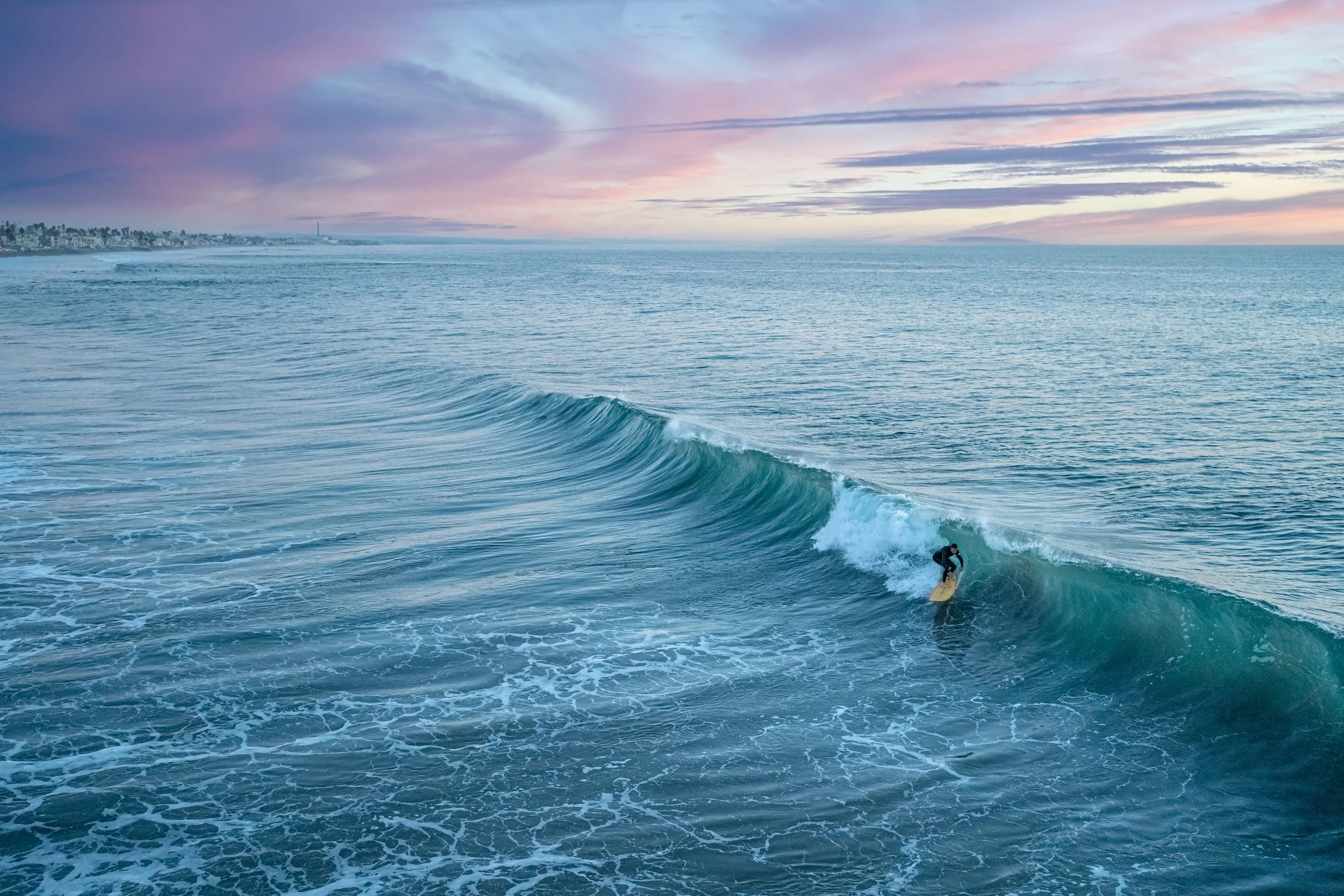 A person surfing on a wave in the ocean during sunset, with a city skyline in the background and colorful sky.