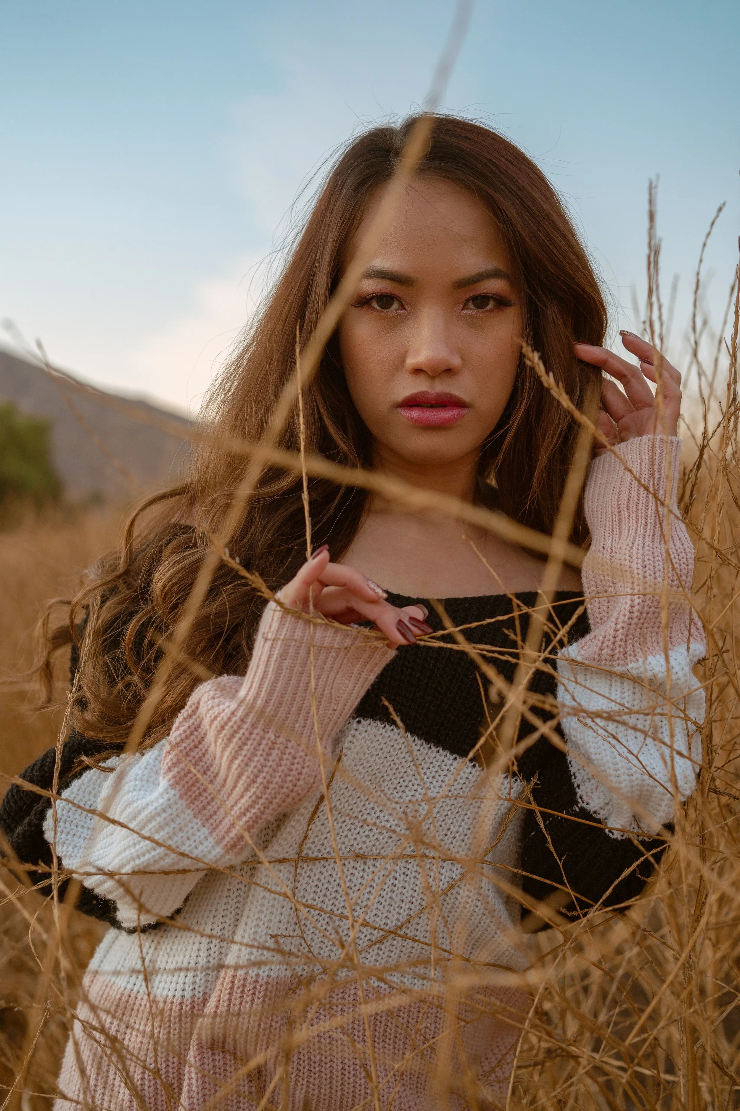 A woman with long brown hair and makeup standing in a field of dry grass, wearing a black top and a beige knitted cardigan, looking directly at the camera with a serious expression.