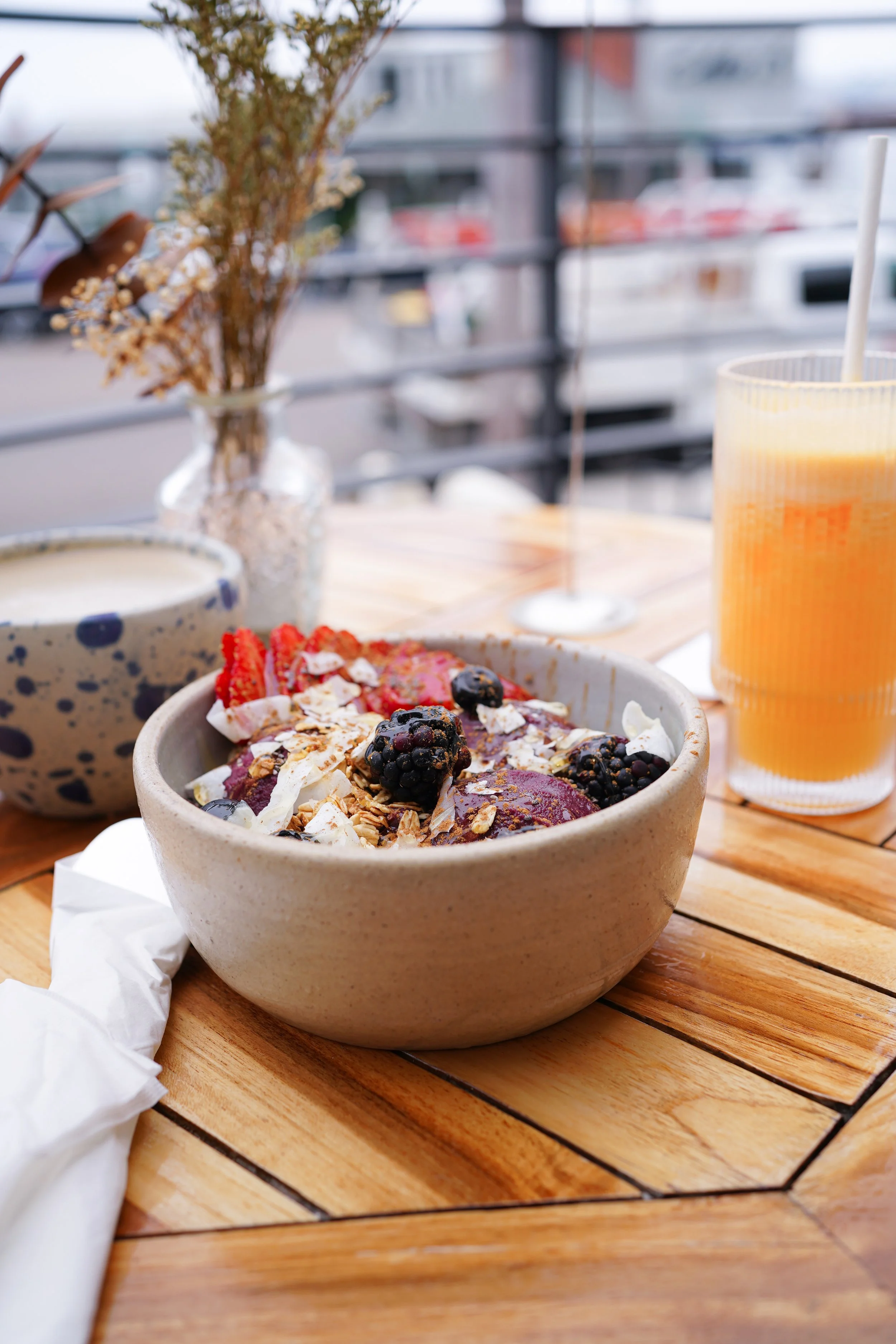 A bowl of yogurt topped with mixed berries and granola on a wooden table, with a glass of orange juice and a cup in the background.