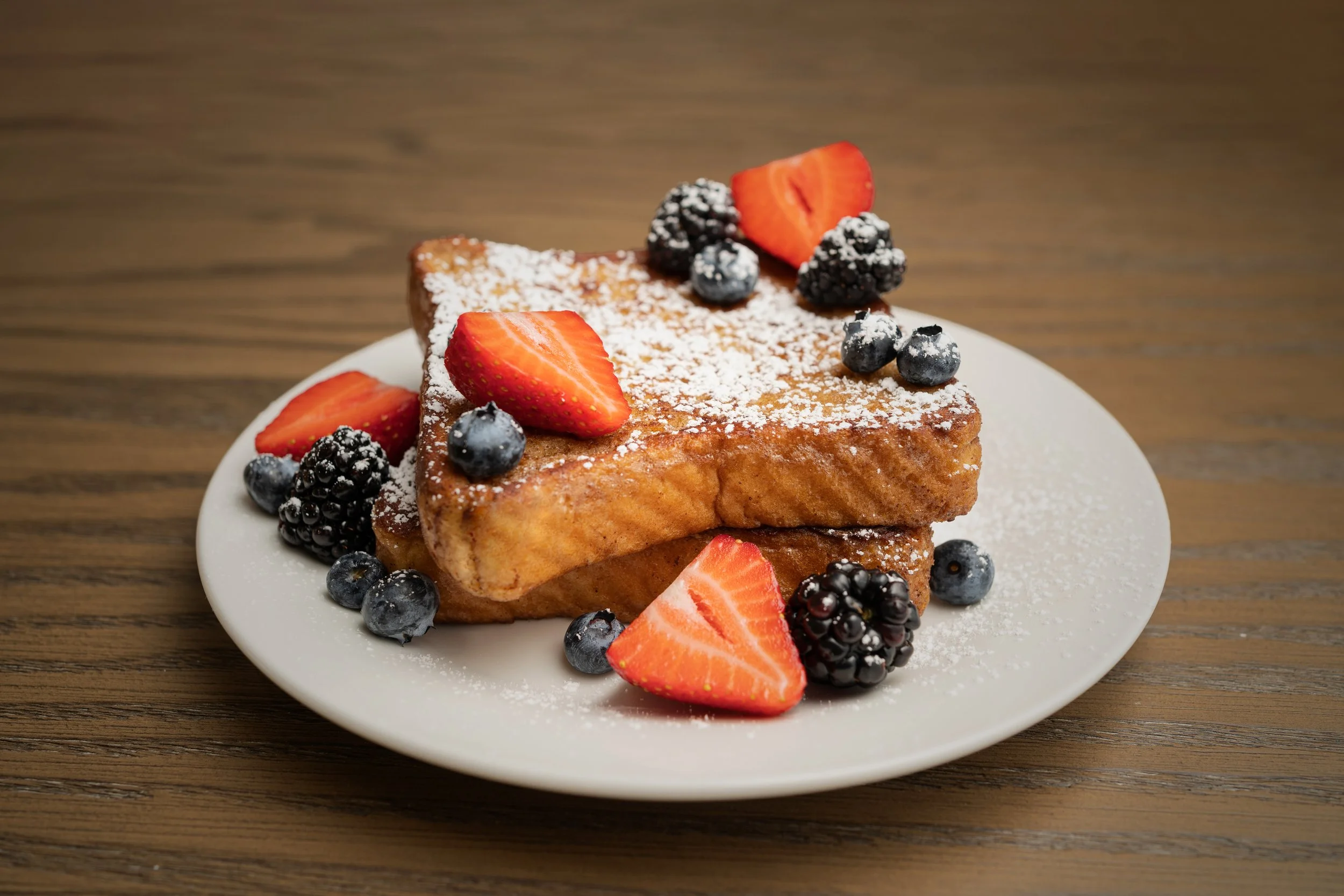 A plate of French toast topped with powdered sugar, sliced strawberries, blueberries, and blackberries on a wooden table.