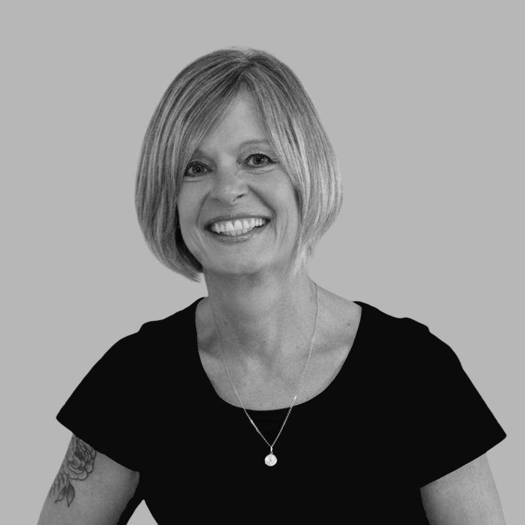 A black and white portrait of a woman with shoulder-length blonde hair, smiling, wearing a black top, a necklace with a circular pendant, and a tattoo on her left arm, against a plain background.