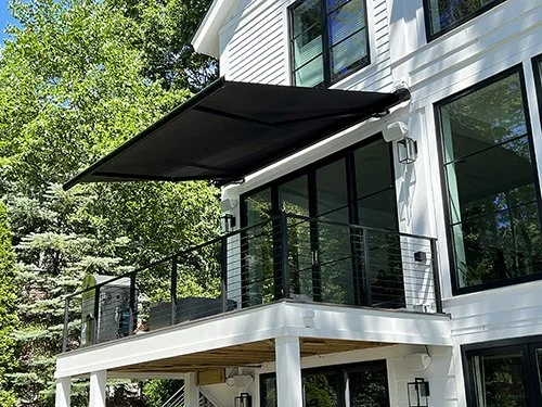 Black Markilux 6000 retractable awning extended over a second-story balcony on a modern white home with large glass doors and black metal railing, surrounded by green trees.