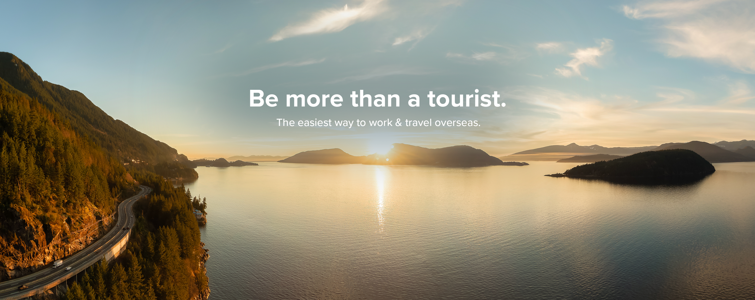 Sunset over a large body of water with surrounding mountains and a road with vehicles on the left side.