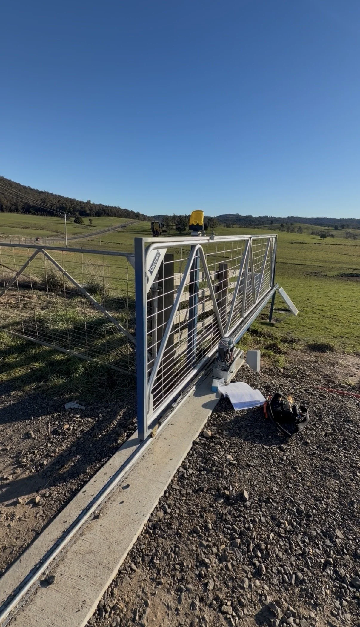 A remote-controlled gate being installed on a rural dirt road with green fields and rolling hills in the background under a clear blue sky.