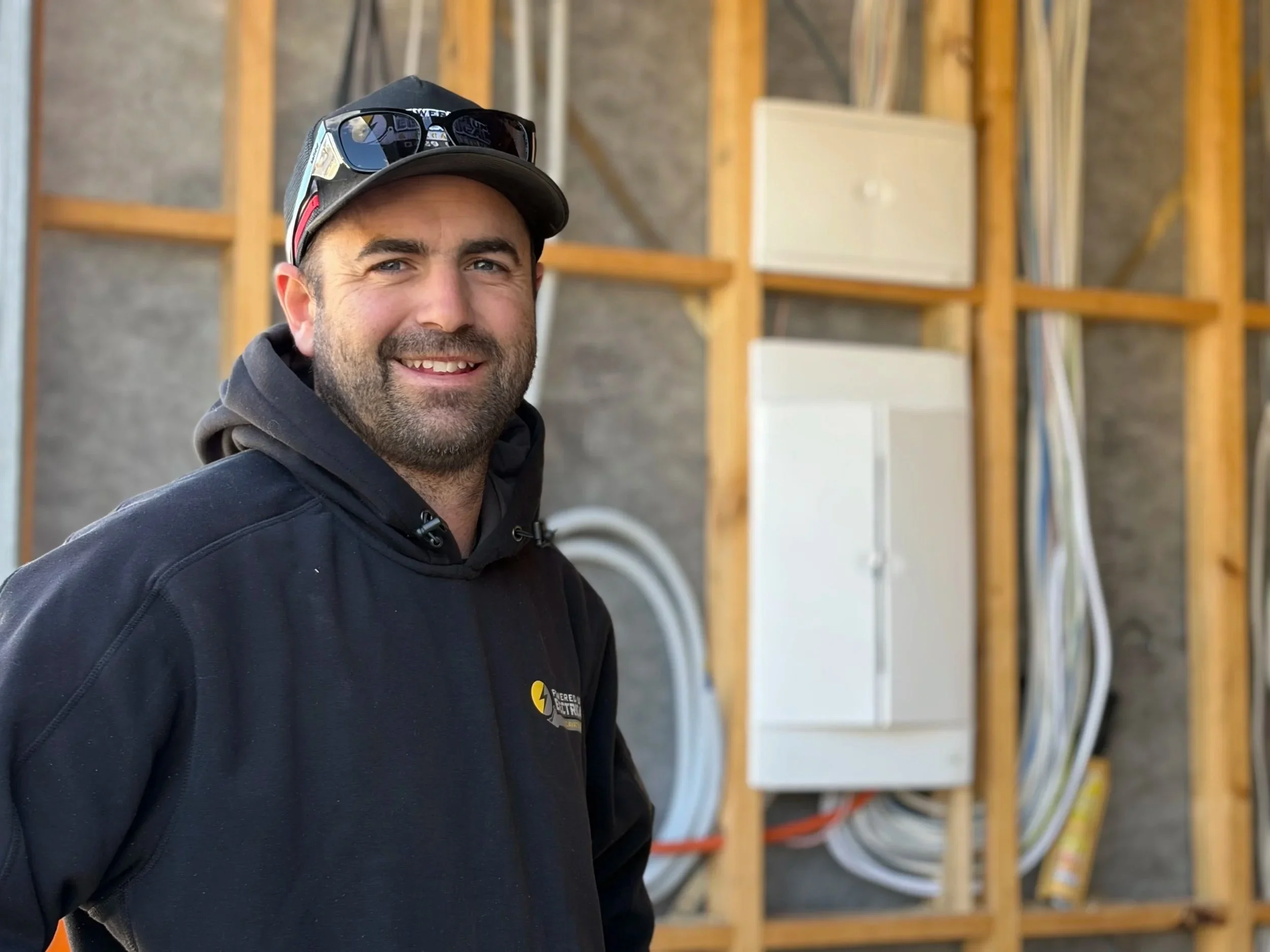 Powered Up Electrical TAS director Joel smiling, wearing a Powered Up Electrical TAS hoodie in front of a switchboard in a new build.