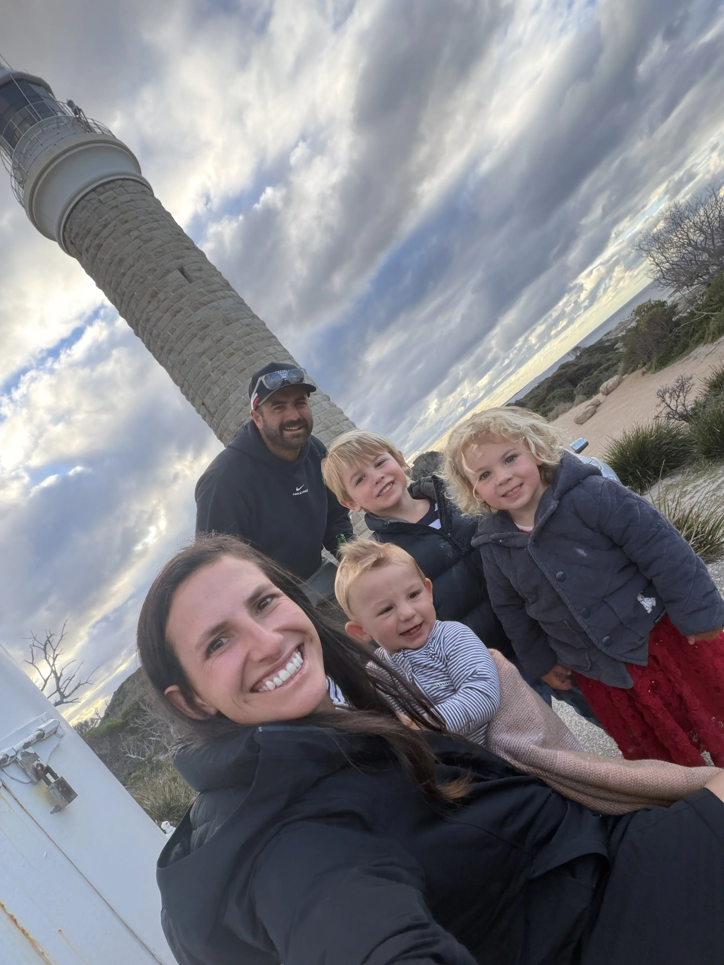 A group of smiling people, including children and adults, taking a selfie with a lighthouse and cloudy sky in the background.