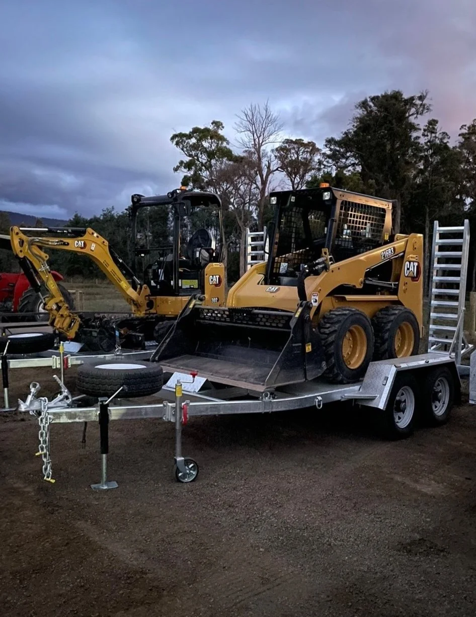 A Caterpillar skid steer and Excavator loaders on trailers, surrounded by trees and cloudy sky.