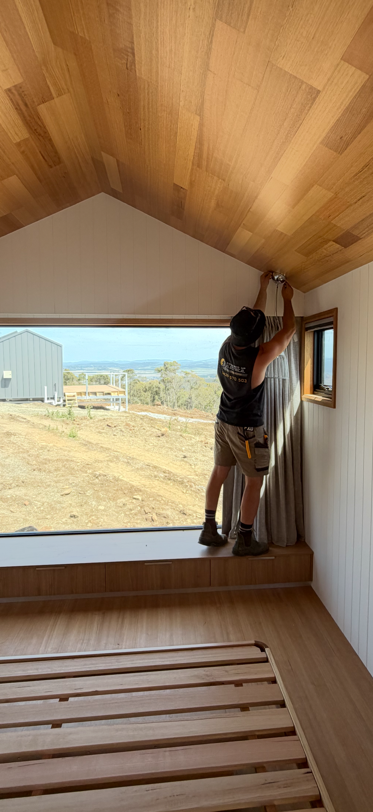 Powered Up Electrical TAS director Joel installing a ceiling light in a room with a large window, wooden ceiling.
