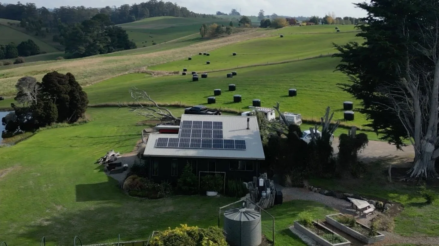 A rural landscape with a house equipped with solar panels on the roof, surrounded by green fields, trees, and hay bales in the distance.