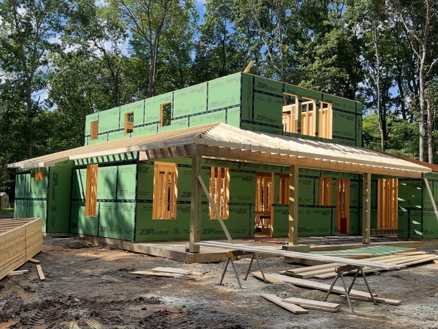 A house under construction in a wooded area, with green insulation panels, wooden framing, and a partially built roof.