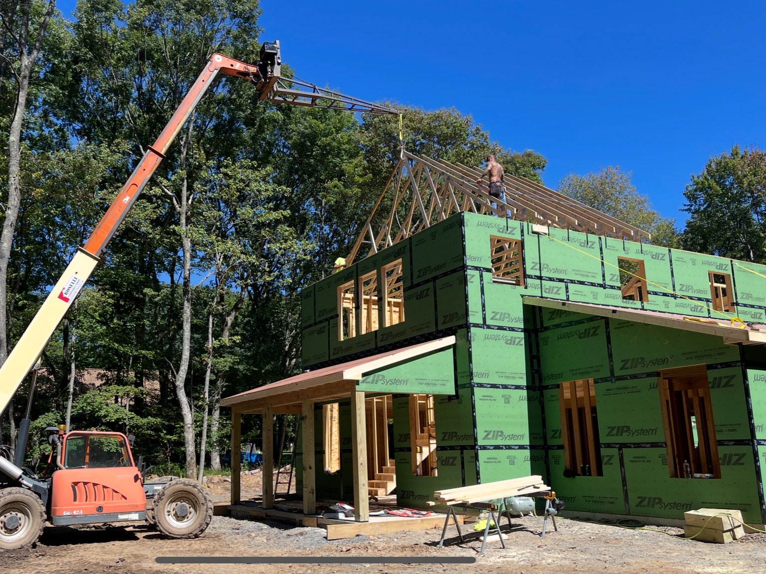 Construction site with a partially built house surrounded by trees, a crane lifting roof trusses, and a worker standing on the roof structure.