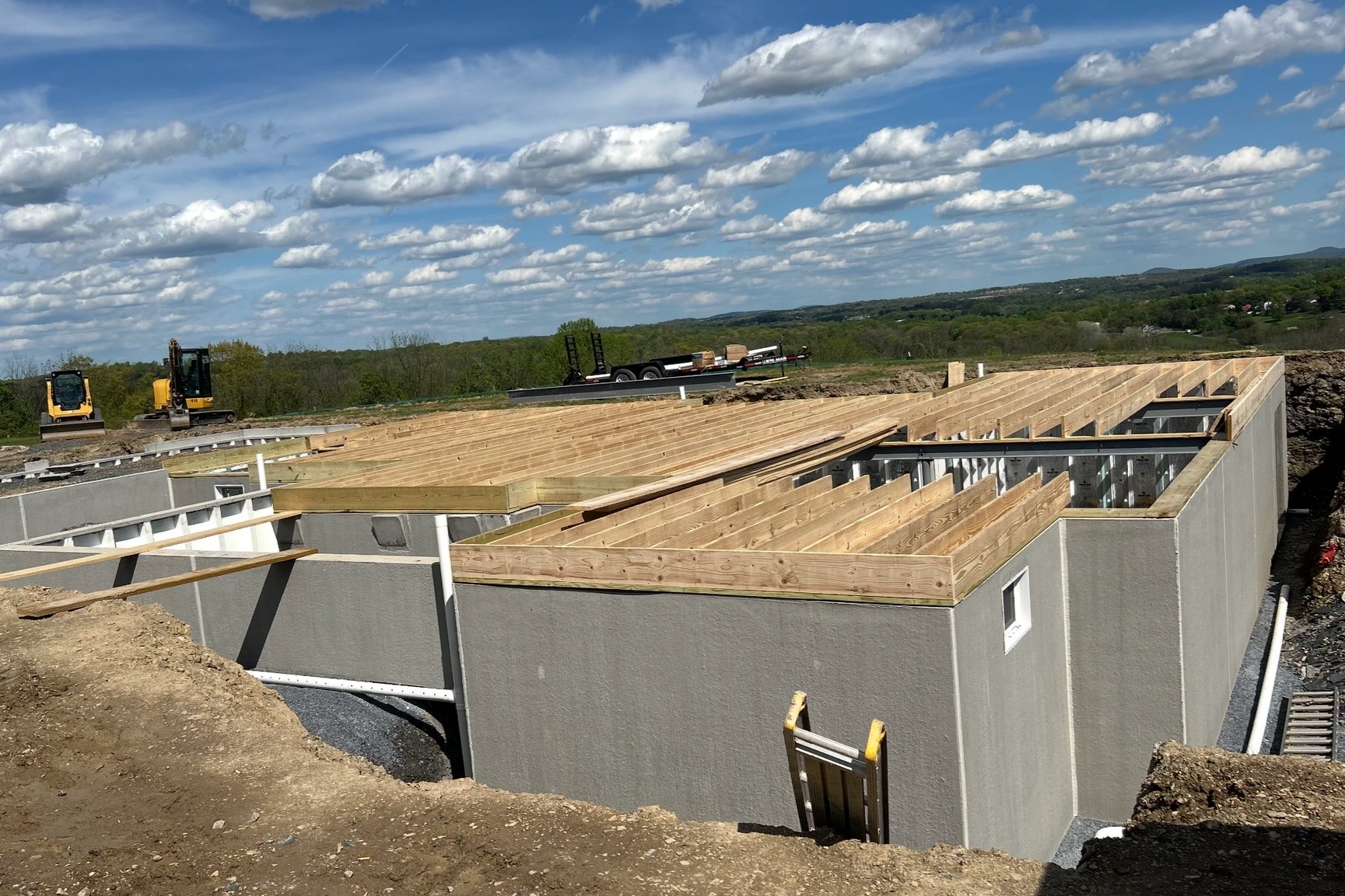 Construction site of a building with wooden roof framework and gray exterior walls, surrounded by dirt and equipment, under a partly cloudy sky.