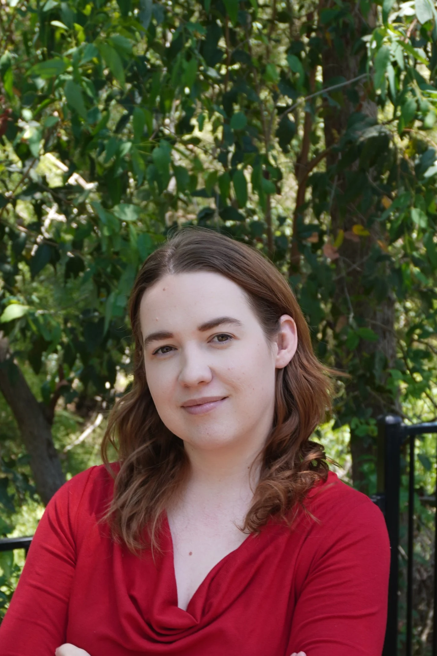 Smiling woman with short brown hair, wearing a black turtleneck and patterned red jacket, sitting in front of a white wall with a wooden shelf holding two potted plants, one tall aloe vera and one fern.