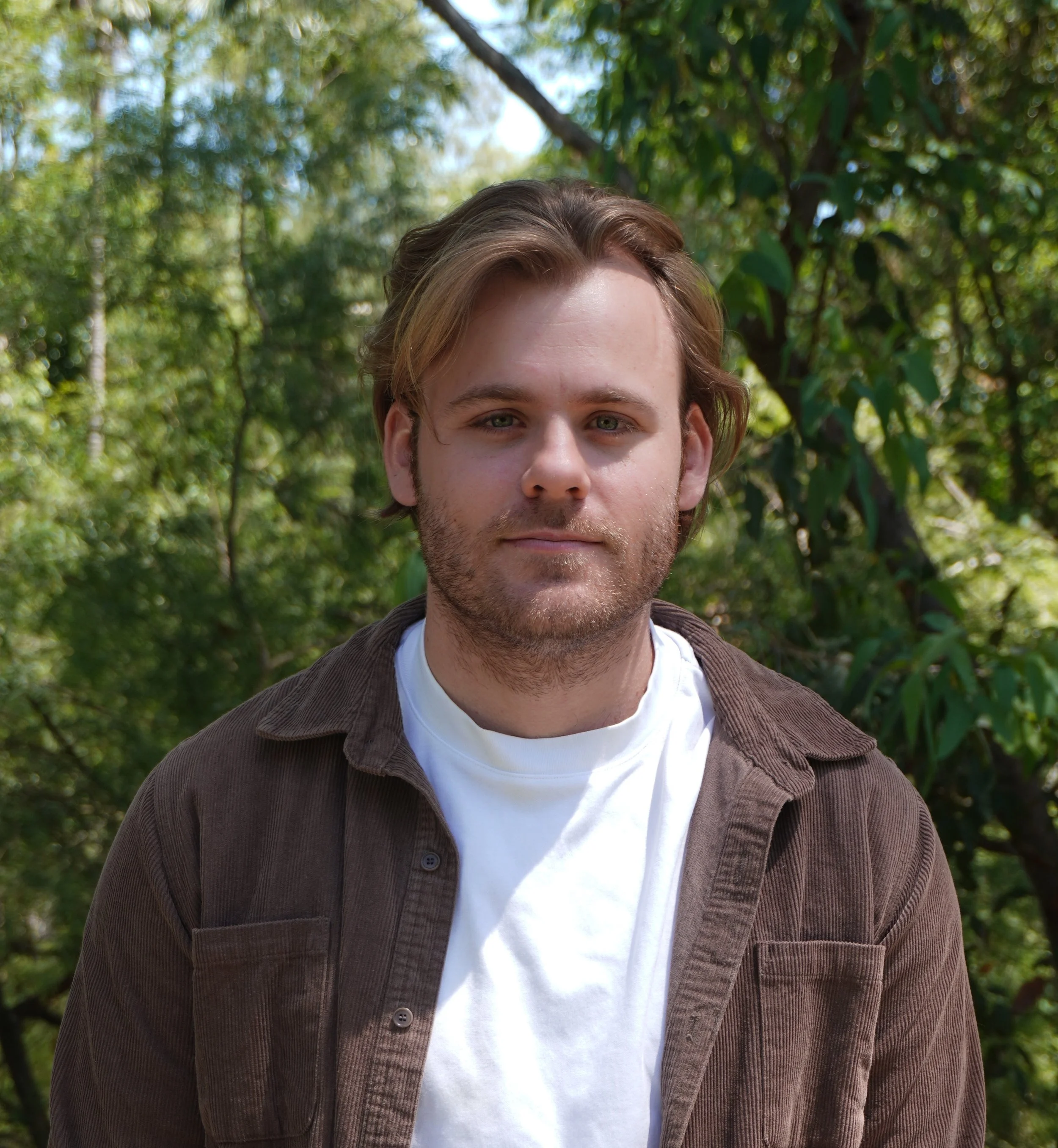 A young man with long hair and a beard standing on a bridge or waterfront in Paris, France, with the Eiffel Tower in the background, near the Seine River with a boat passing by.
