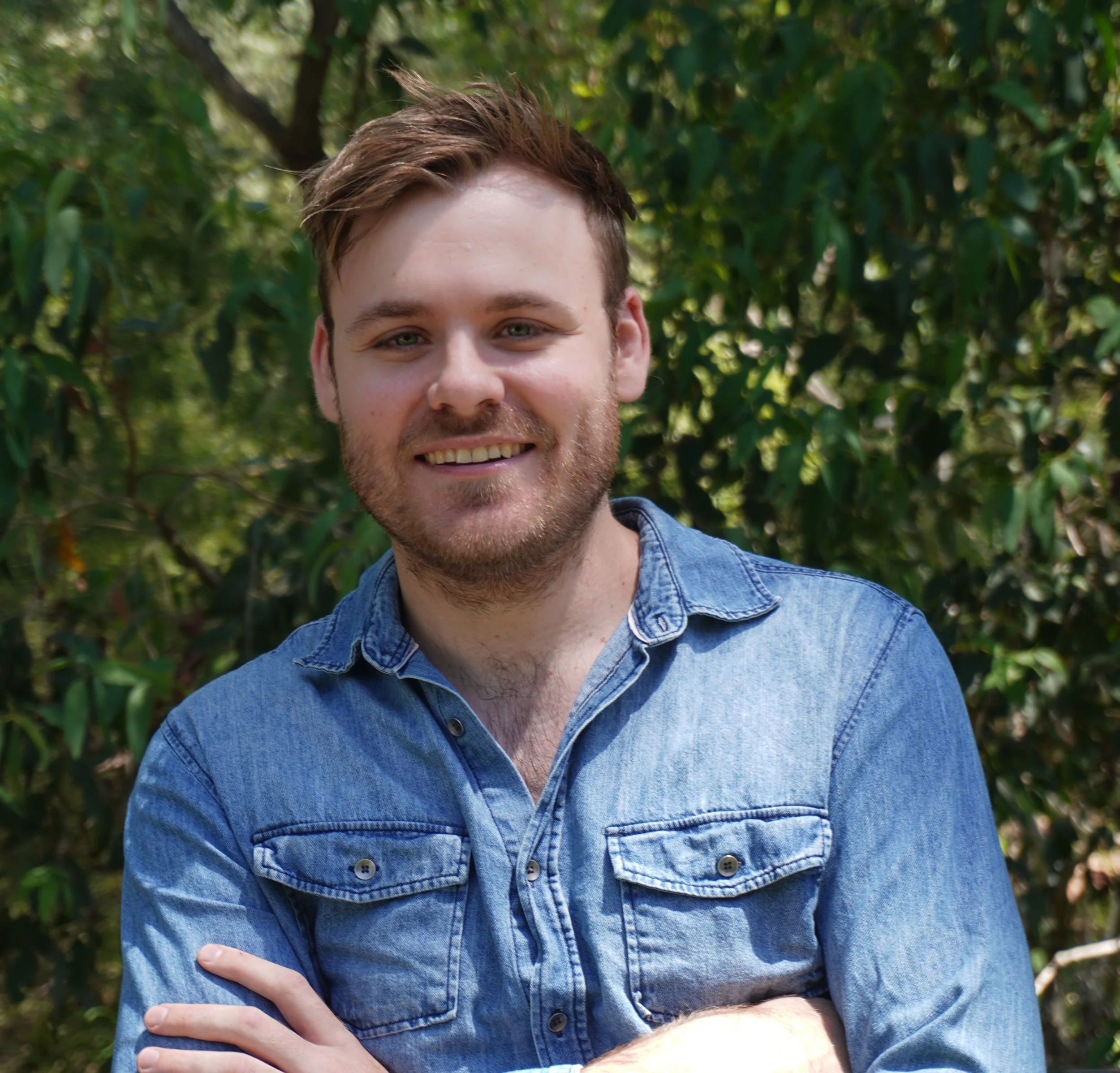 Man wearing sunglasses, a white beanie, and a white sweater standing in snowy mountains, making a peace sign with one hand, holding a jacket in the other, with snow-covered peaks and a blue sky in the background.