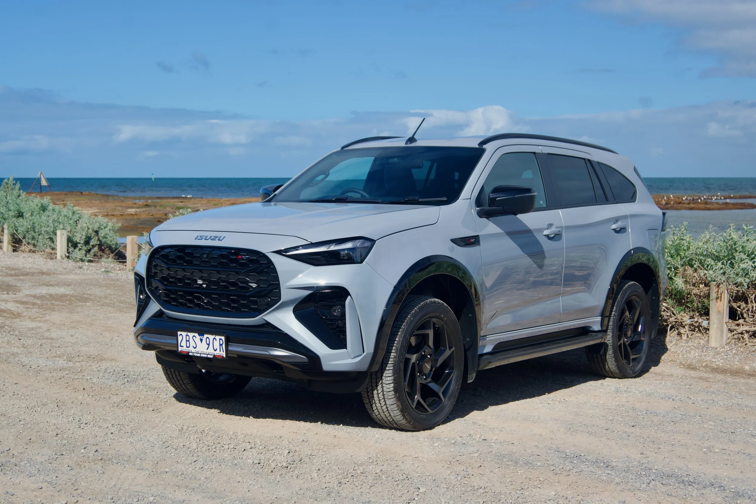 A silver Isuzu SUV parked near the beach with the ocean and blue sky in the background.