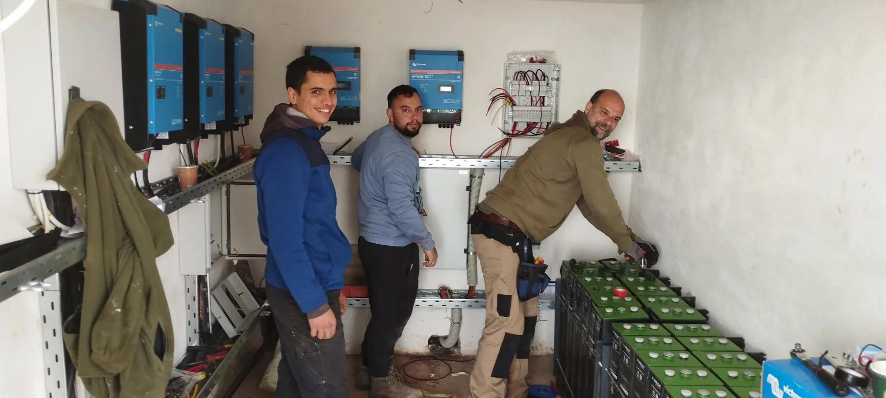 Three men working in an electrical room with batteries and electrical panels.
