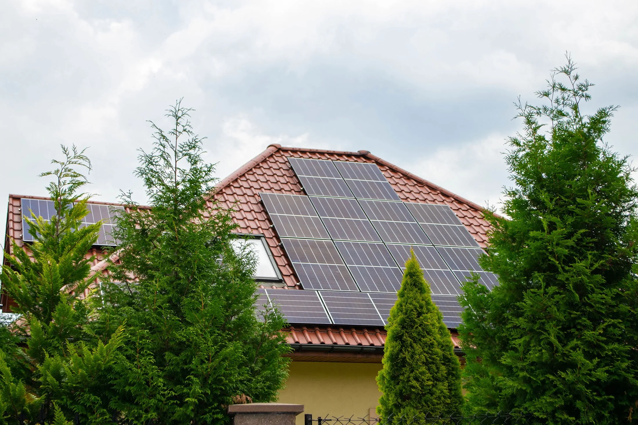 House with solar panels on a red-tiled roof surrounded by green trees.