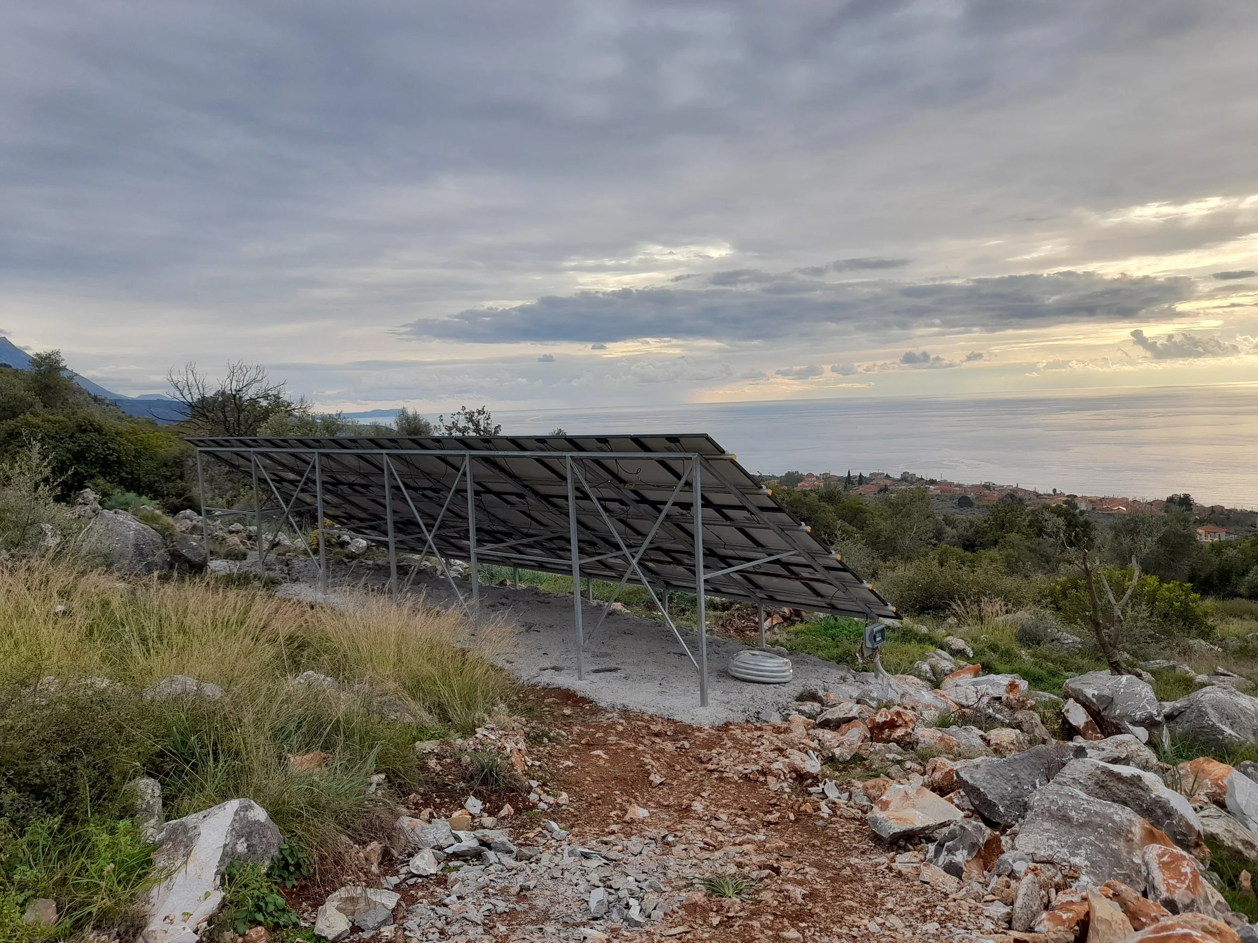 Solar panel array installed on a hillside with a view of the ocean and a cloudy sky in the background.
