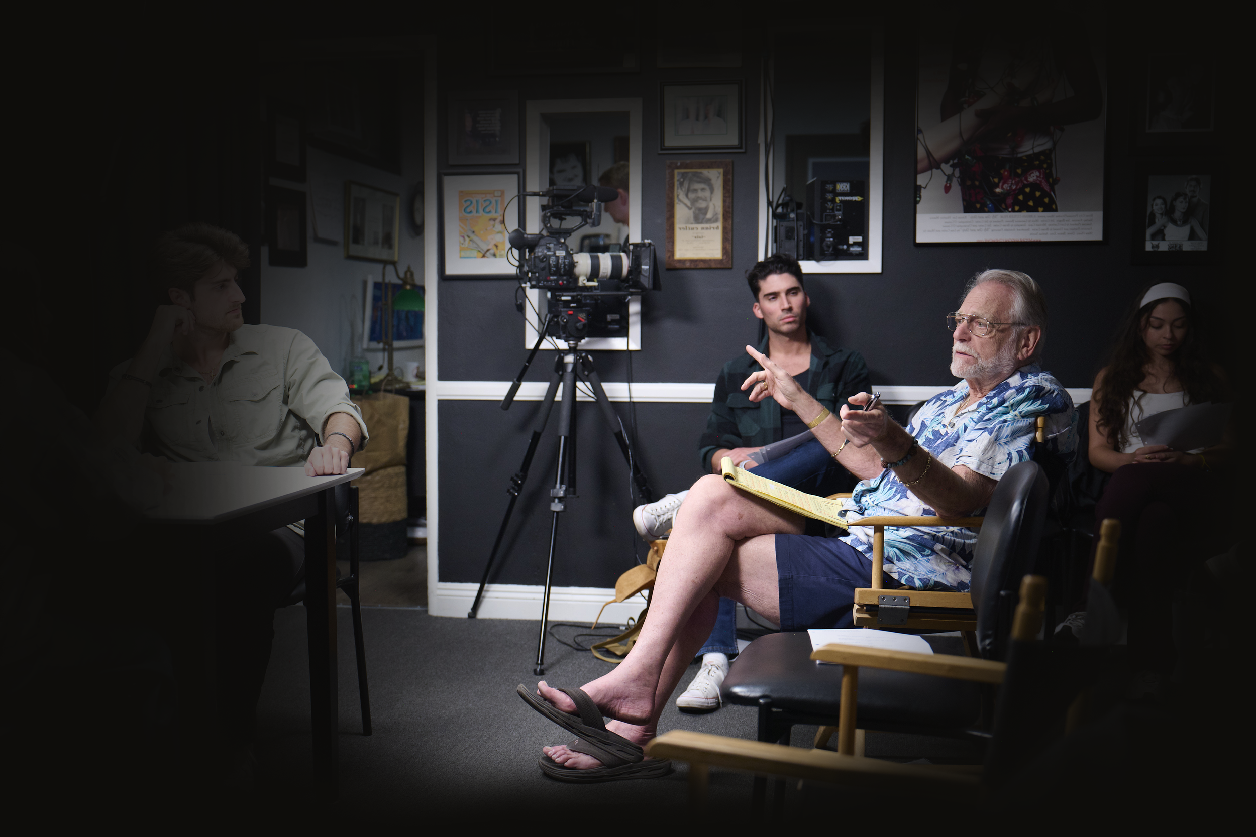 A film crew seated in a black-walled room with photos on the walls, a professional camera on a tripod, and crew members, including Brian Cutler in Hawaiian shirt and shorts holding a notepad and talking, while others listen or look at devices.